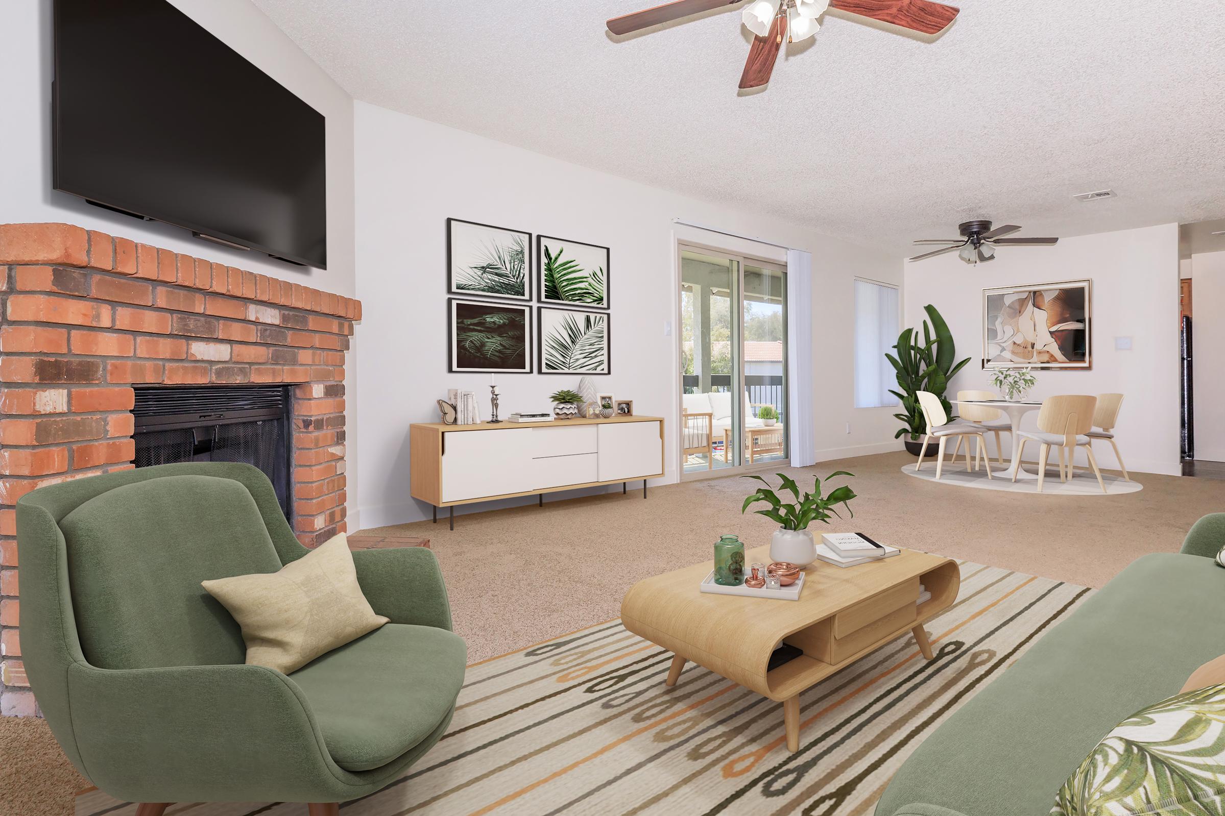 Cozy living room featuring a green armchair, wooden coffee table, and a brick fireplace. The decor includes framed botanical art on the wall, a modern TV mounted above the fireplace, and a bright atmosphere with sliding glass doors leading to an outdoor area. Light-colored walls and a soft rug complete the inviting space.