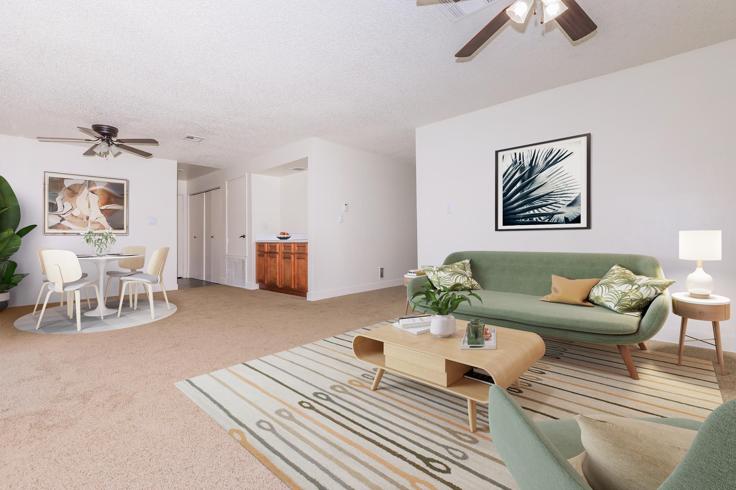 A modern living room featuring a green sofa, a wooden coffee table, and a patterned rug. There is a small dining area with white chairs and a round table. The walls are light-colored, with a framed leaf print on one wall. A ceiling fan adds to the ambiance, and there's an indoor plant in the corner.