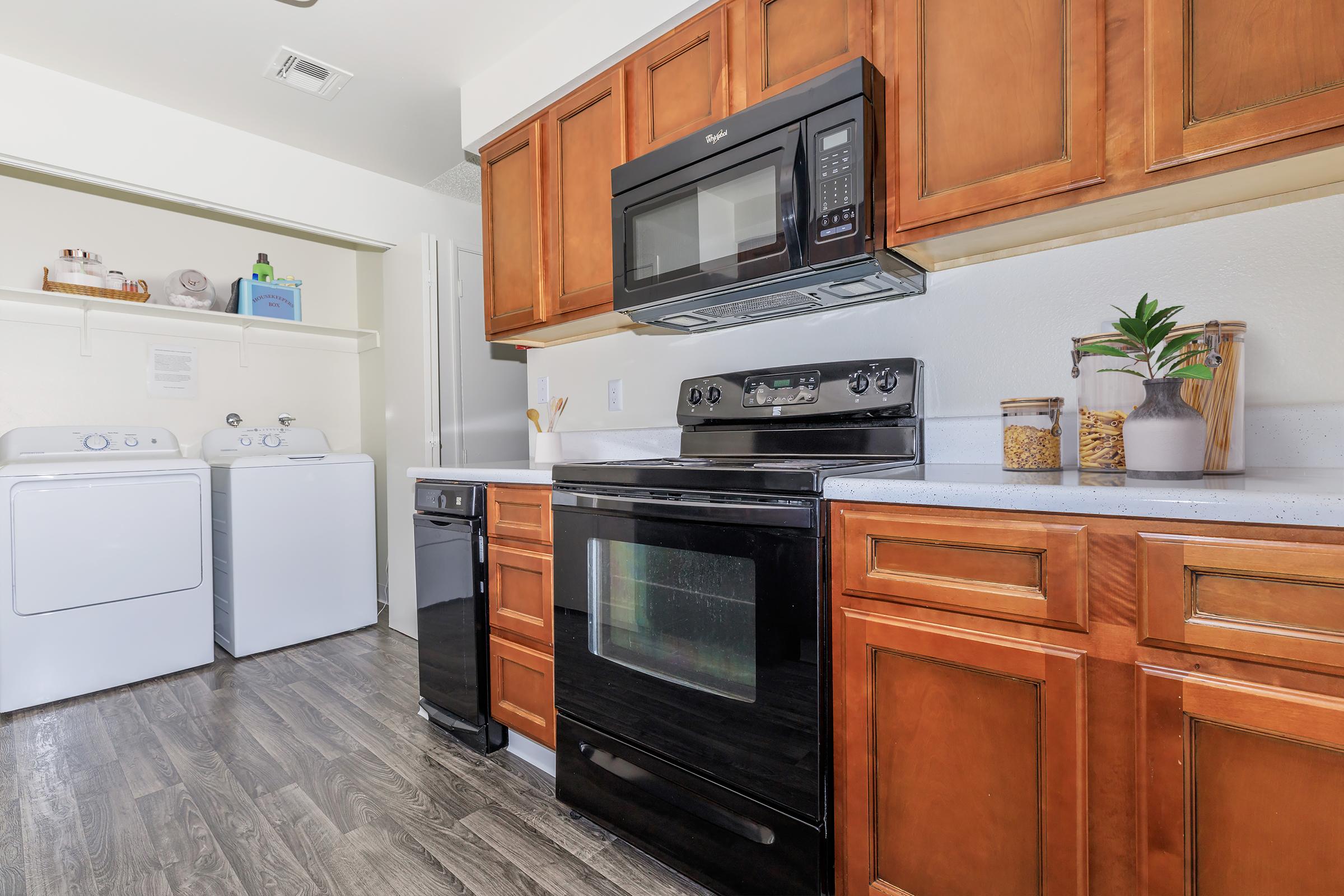 A modern kitchen featuring wooden cabinets, a black stove, microwave, and dishwasher. In the background, there is a laundry area with a washing machine and dryer. The countertop has decorative items including a plant and a jar, and the flooring is a dark wood laminate.