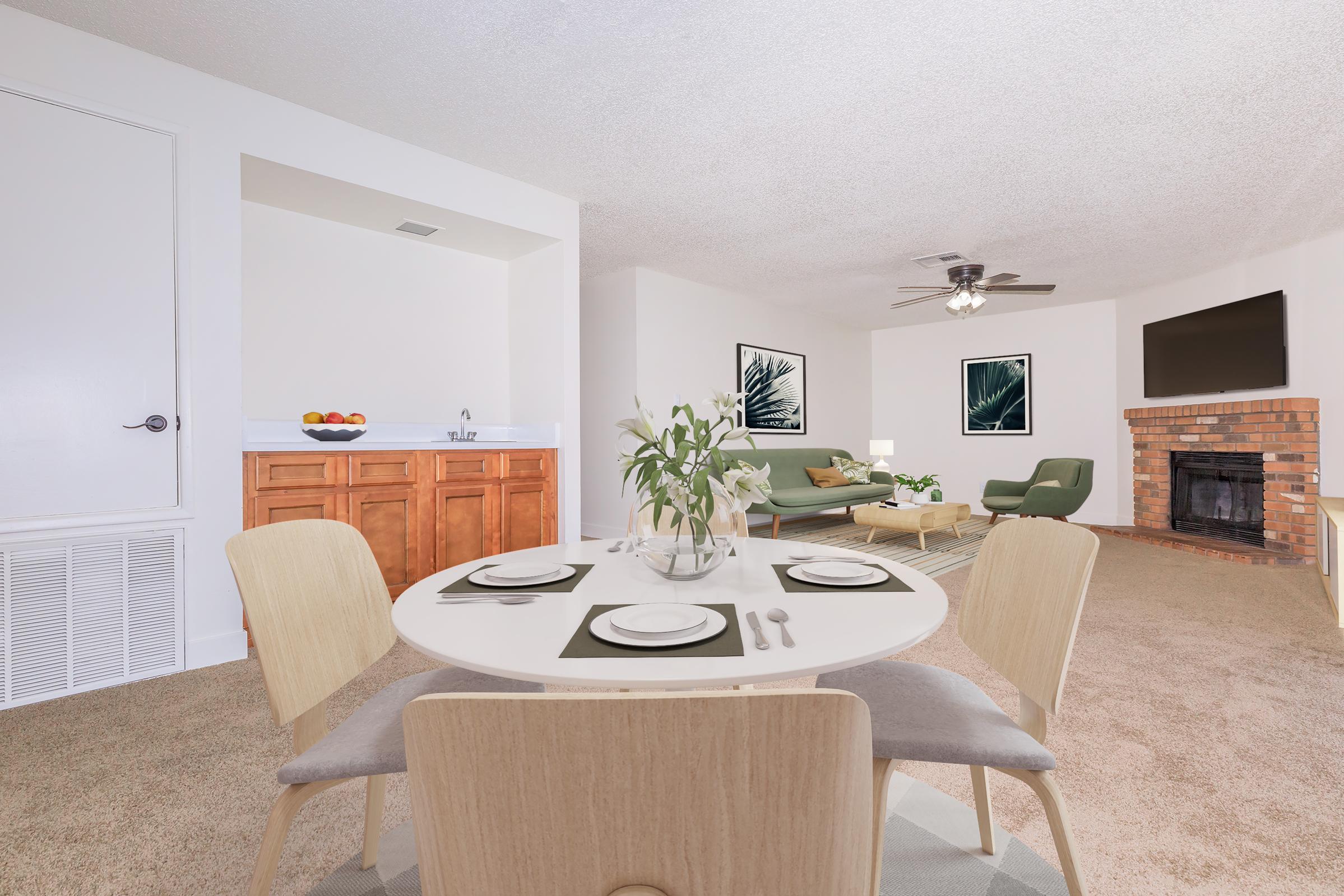 A cozy living and dining area featuring a round dining table set for four, with a vase of flowers in the center. The space includes a light-filled living room with a green sofa, a brick fireplace, and framed artwork on the walls. A kitchenette is visible in the background, adding to the inviting atmosphere.