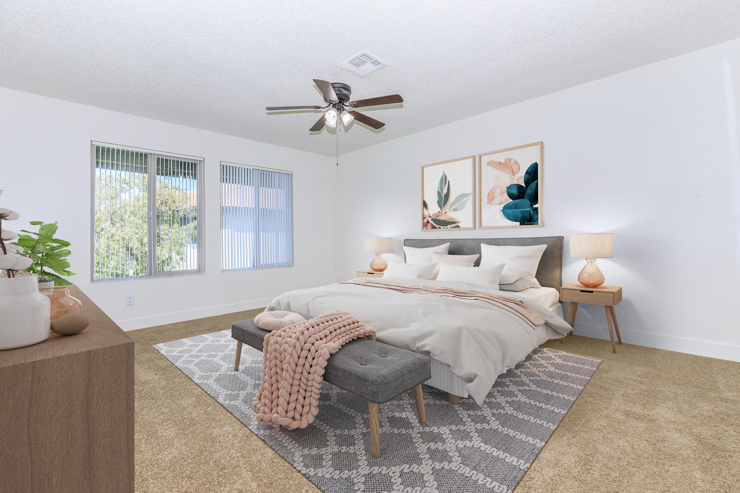 A cozy, well-decorated bedroom featuring a large bed with soft bedding, two bedside lamps on wooden tables, and a textured gray rug. The room has three windows letting in natural light, and two framed artworks with botanical themes hang above the bed. A stylish plant adds a touch of greenery.