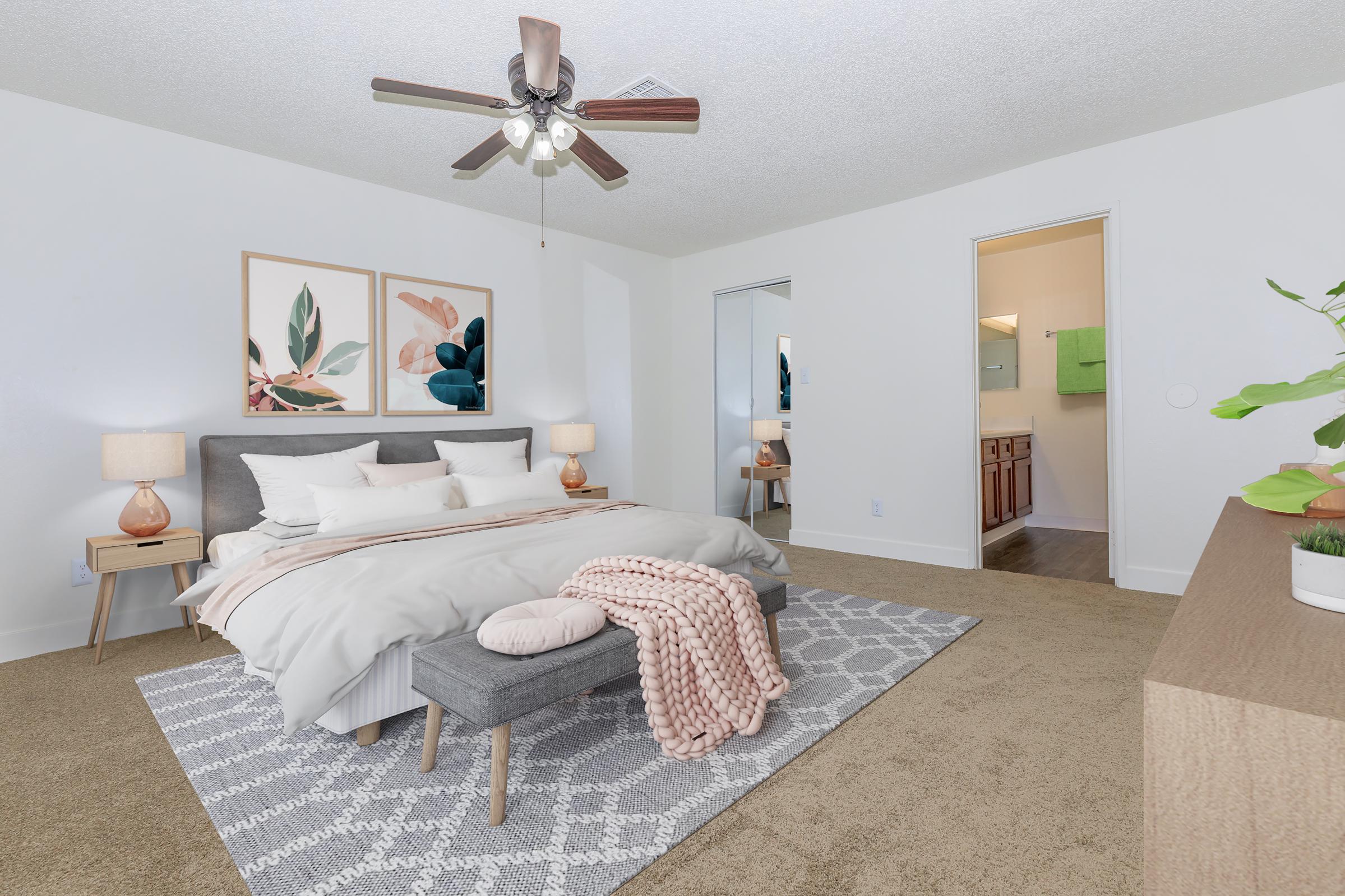 A spacious, modern bedroom featuring a large bed with white bedding and decorative pillows, a gray ottoman at the foot, two stylish lamps on nightstands, and framed botanical art on the walls. There's a large mirror and an open door leading to a bathroom, with light-colored walls and a cozy carpet.