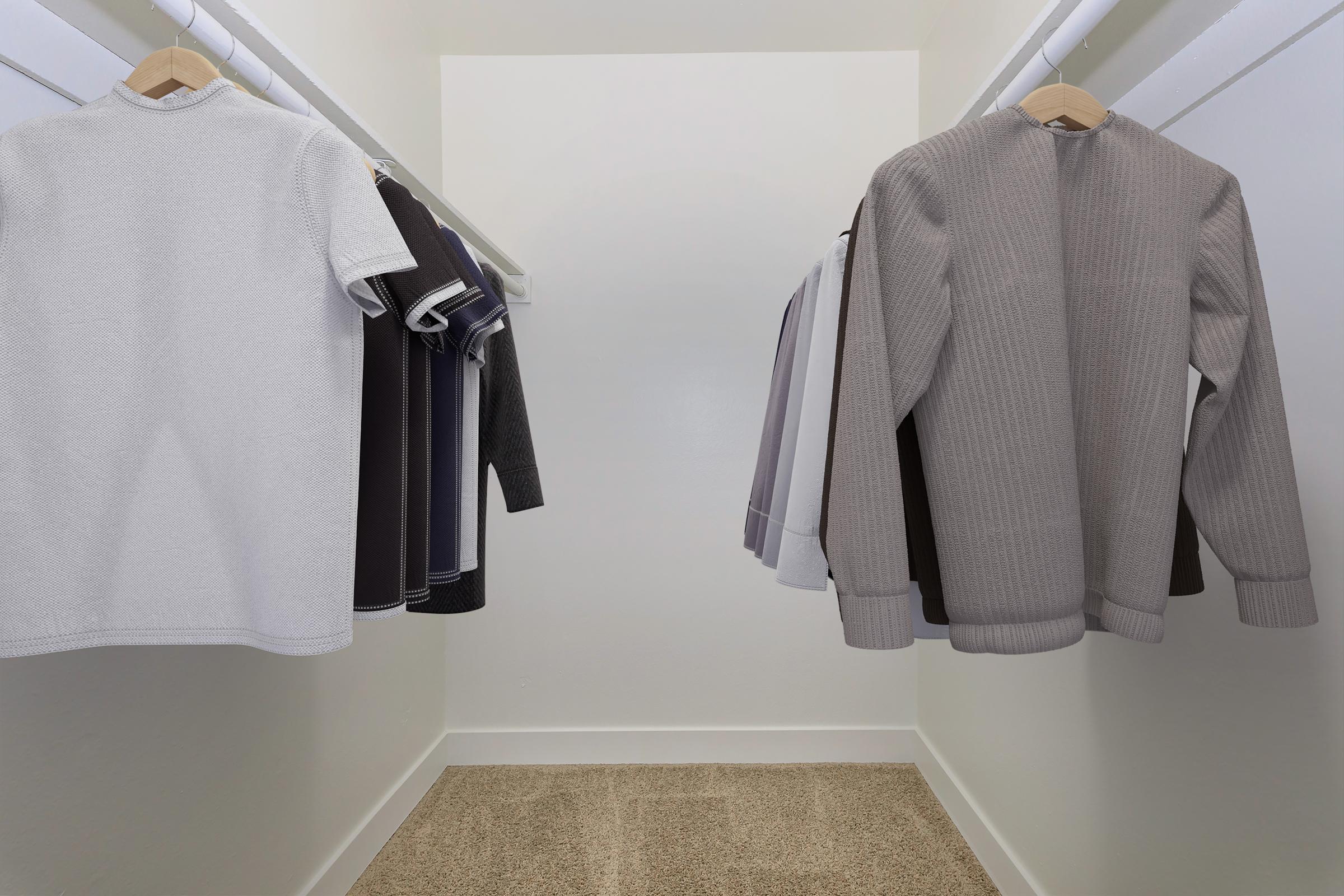 A well-organized closet featuring several hanging shirts in neutral tones, including white, gray, and dark colors, with a light carpeted floor and plain white walls, creating a minimalist and tidy aesthetic.