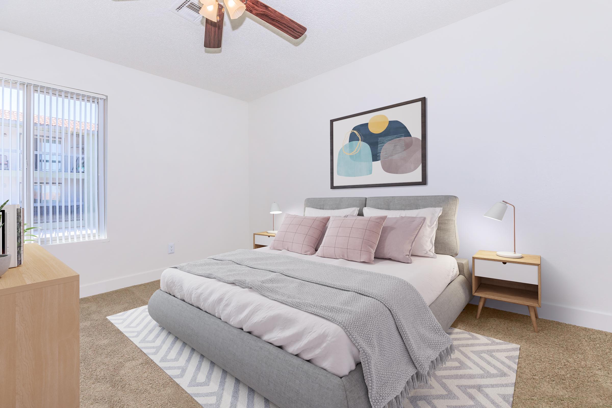 A cozy bedroom featuring a queen-sized bed with light gray bedding and decorative pillows, a ceiling fan, two bedside lamps on wooden tables, and a minimalist wall art piece. The room has light-colored walls, a plush carpet, and natural light coming through a window with blinds.
