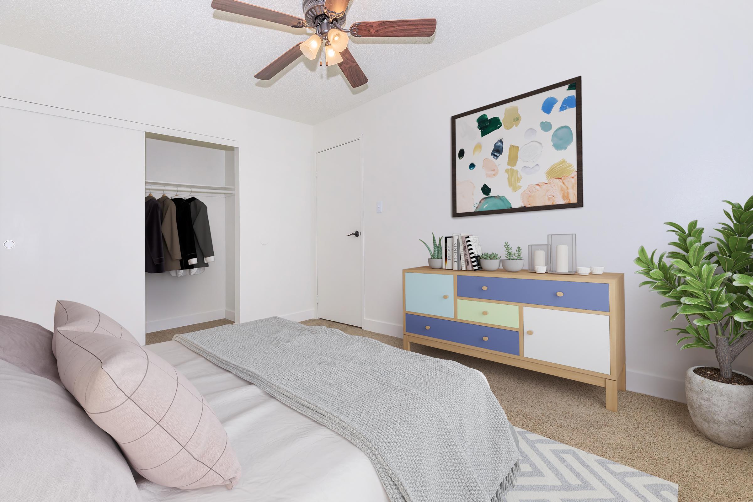 A cozy bedroom featuring a neatly made bed with gray and pink bedding. A colorful abstract painting hangs on the wall above a stylish dresser with blue and white drawers. A small indoor plant and a decorative candle sit on the dresser, while a closet with hanging clothes is visible in the background. A ceiling fan adds to the ambiance.