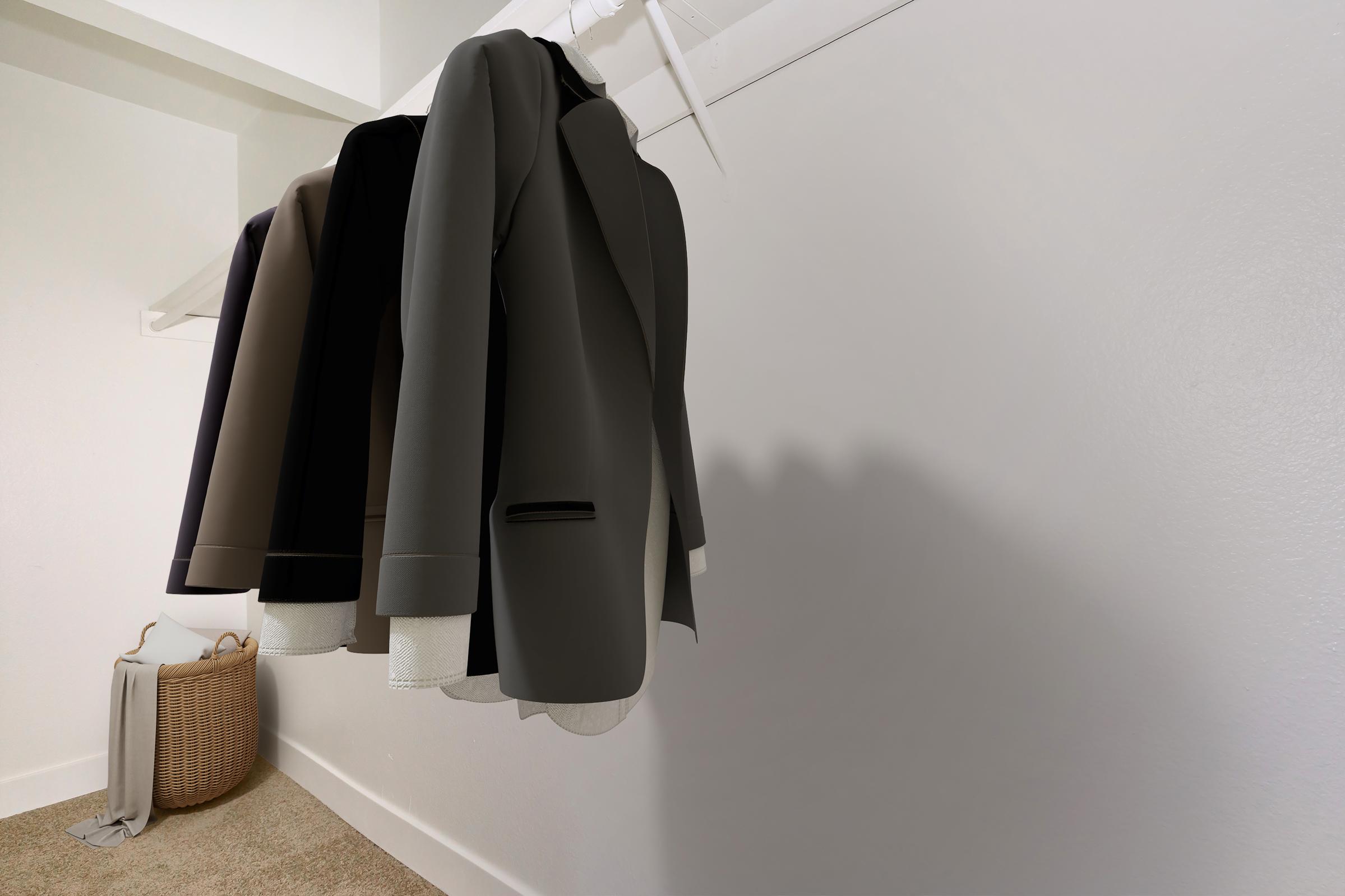 Three suits hanging on a white rack against a light-colored wall. The suits are in shades of gray, black, and brown, with visible details like lapels and buttons. In the foreground, a woven basket and a folded fabric are placed on the floor, adding a touch of decor to the minimalist space.
