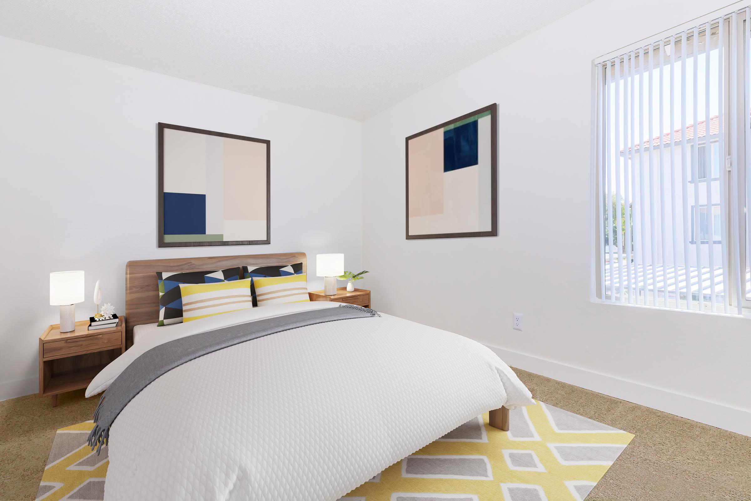 A bright, modern bedroom featuring a bed with patterned yellow and gray bedding. There are two minimalist artworks on the walls, bedside tables with lamps, and a window with vertical blinds letting in natural light. The room has beige carpeting and a geometric area rug.