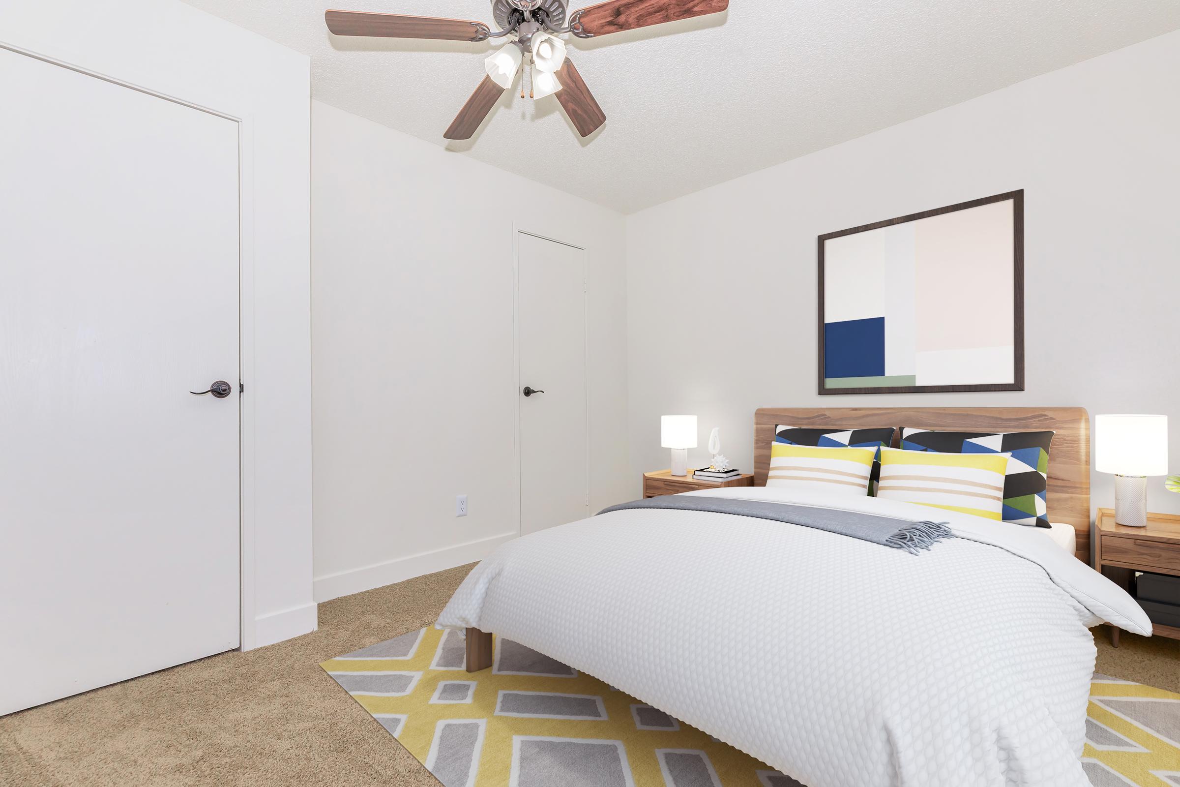 A cozy bedroom featuring a light-colored wall and carpet, a modern wooden bed with a white textured blanket, and colorful striped pillows. There's a bedside table with lamps on either side, and a geometric area rug on the floor. A ceiling fan and a closed door are visible in the background.