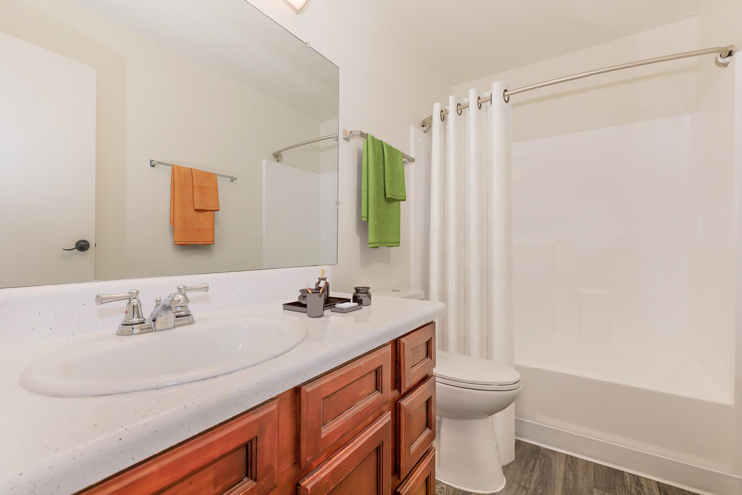 A clean and modern bathroom featuring a white bathtub and shower combination, a light-colored countertop with dual sinks, wooden cabinetry, and a large mirror. Brightly colored towels hang on the towel racks—one orange and one green—while a neutral color palette enhances the space's inviting look.