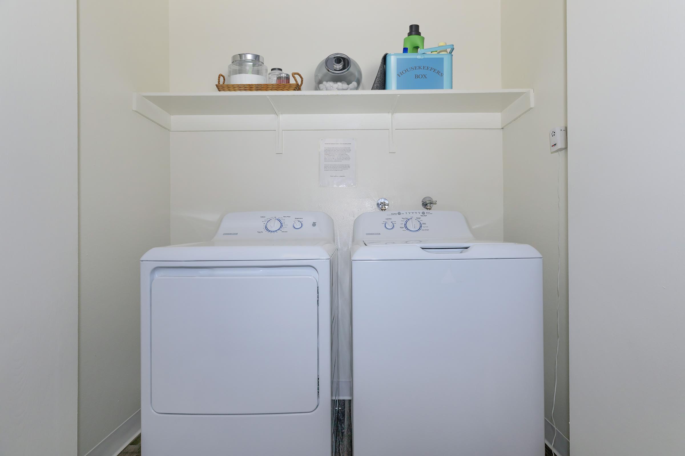 A laundry room with a white washing machine and a dryer side by side. Above them, a shelf holds cleaning supplies, including a basket and a blue container labeled "Laundry." The walls are light-colored, creating a clean and organized appearance.