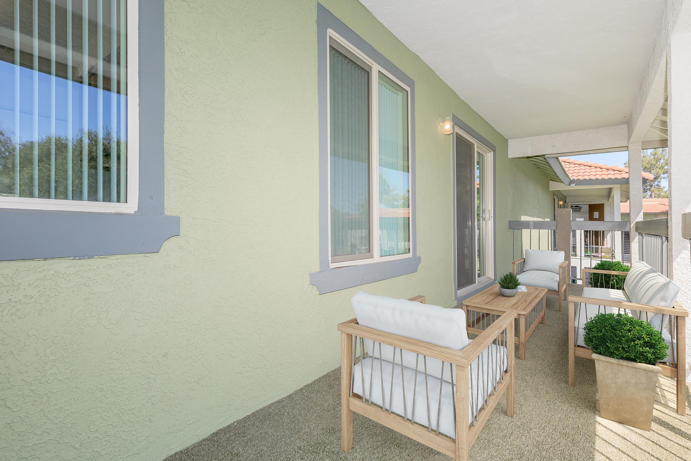 A cozy balcony area featuring light-colored furniture with cushions, small potted plants, and a textured wall painted in a soft green. Natural light streams in through large windows, creating a welcoming outdoor space that's perfect for relaxing or enjoying a morning coffee.
