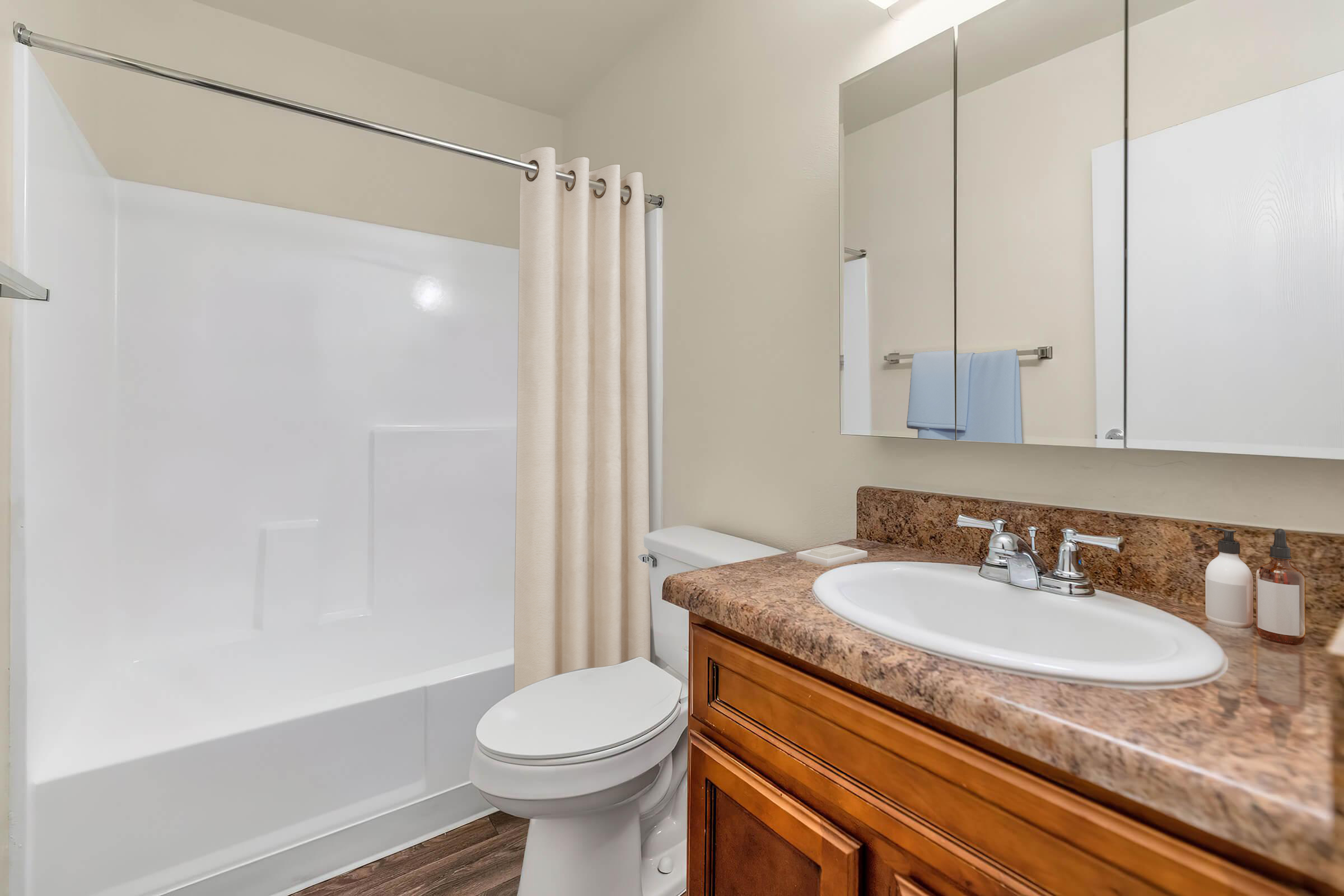 A clean, modern bathroom featuring a white bathtub with a shower curtain, a wooden vanity with a round sink, a toilet, and a large mirror above the sink. The walls are a neutral color, and the flooring appears to be a wood-like material. A blue towel is hanging on a towel rack.