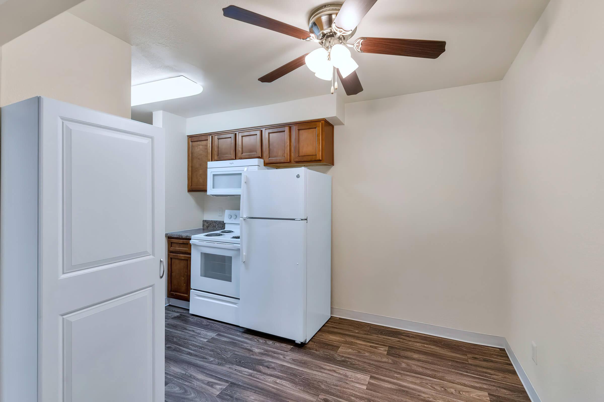 A modern kitchen featuring wooden cabinets, a white refrigerator, and a white oven with a microwave above. The floor is made of dark wood, and there's a ceiling fan with lights. The walls are painted a light color, creating a bright and open atmosphere.