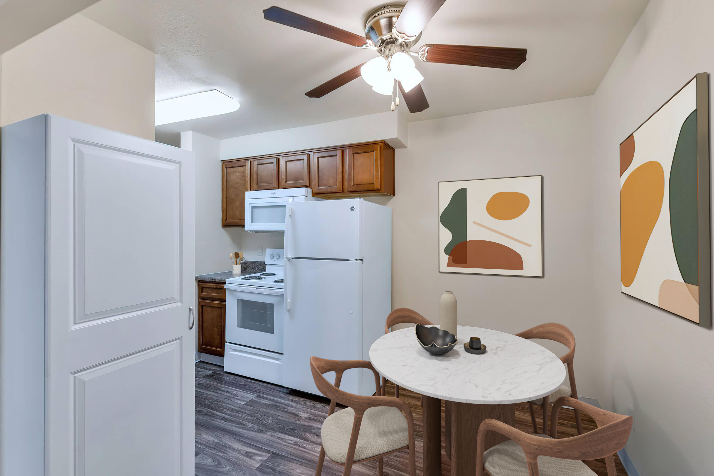 A modern kitchen featuring wooden cabinets, a white stove and fridge, and a ceiling fan. A round marble table with stylish chairs is set up for dining, accompanied by abstract artwork on the walls. The space is bright and inviting, showcasing a blend of contemporary design elements.