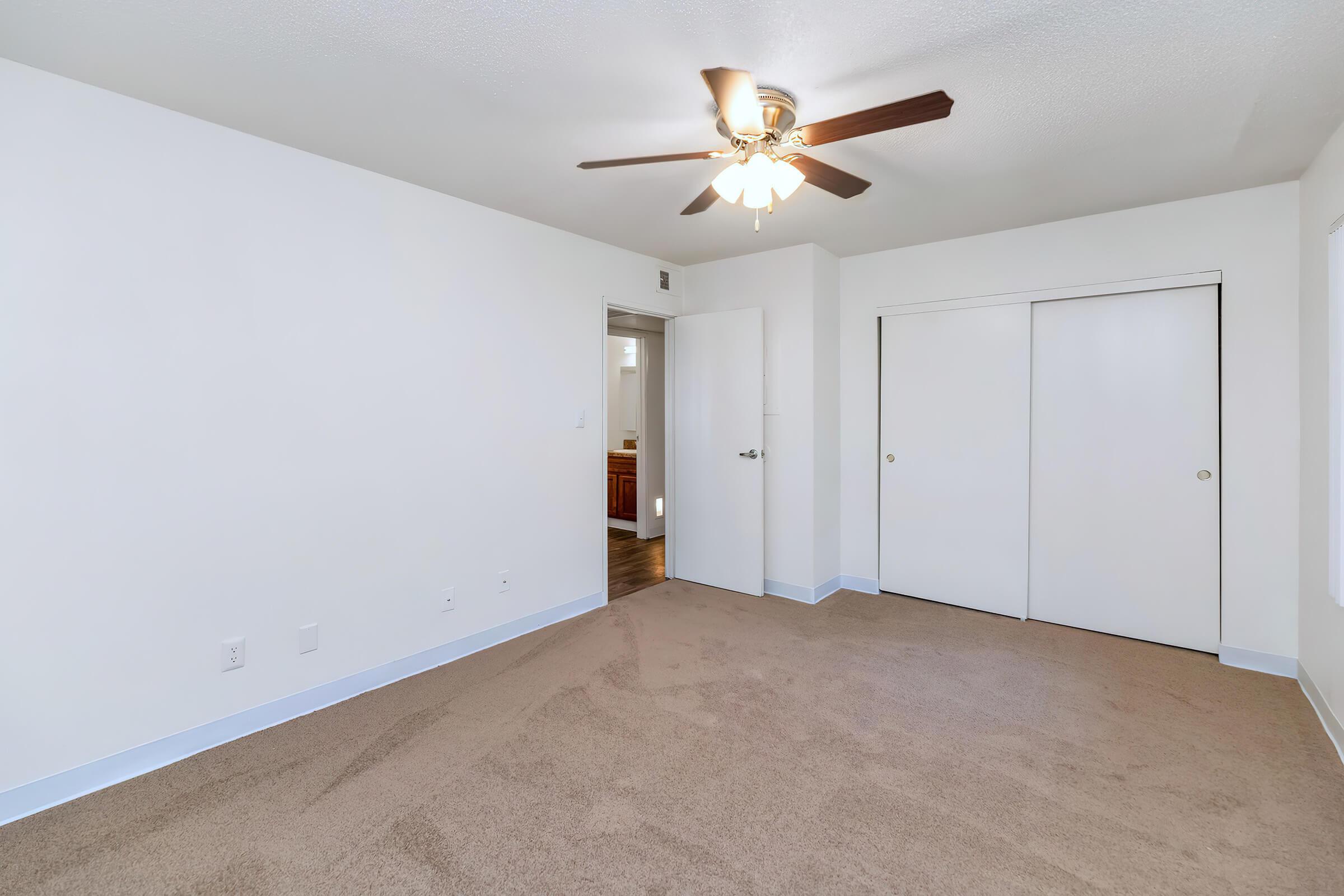 A spacious, empty room featuring light-colored walls and soft, beige carpet. A ceiling fan with wooden blades hangs from the ceiling. There are two doors: one leading to a closet and the other to an adjoining space. Natural light enters through a window, creating a bright and inviting atmosphere.