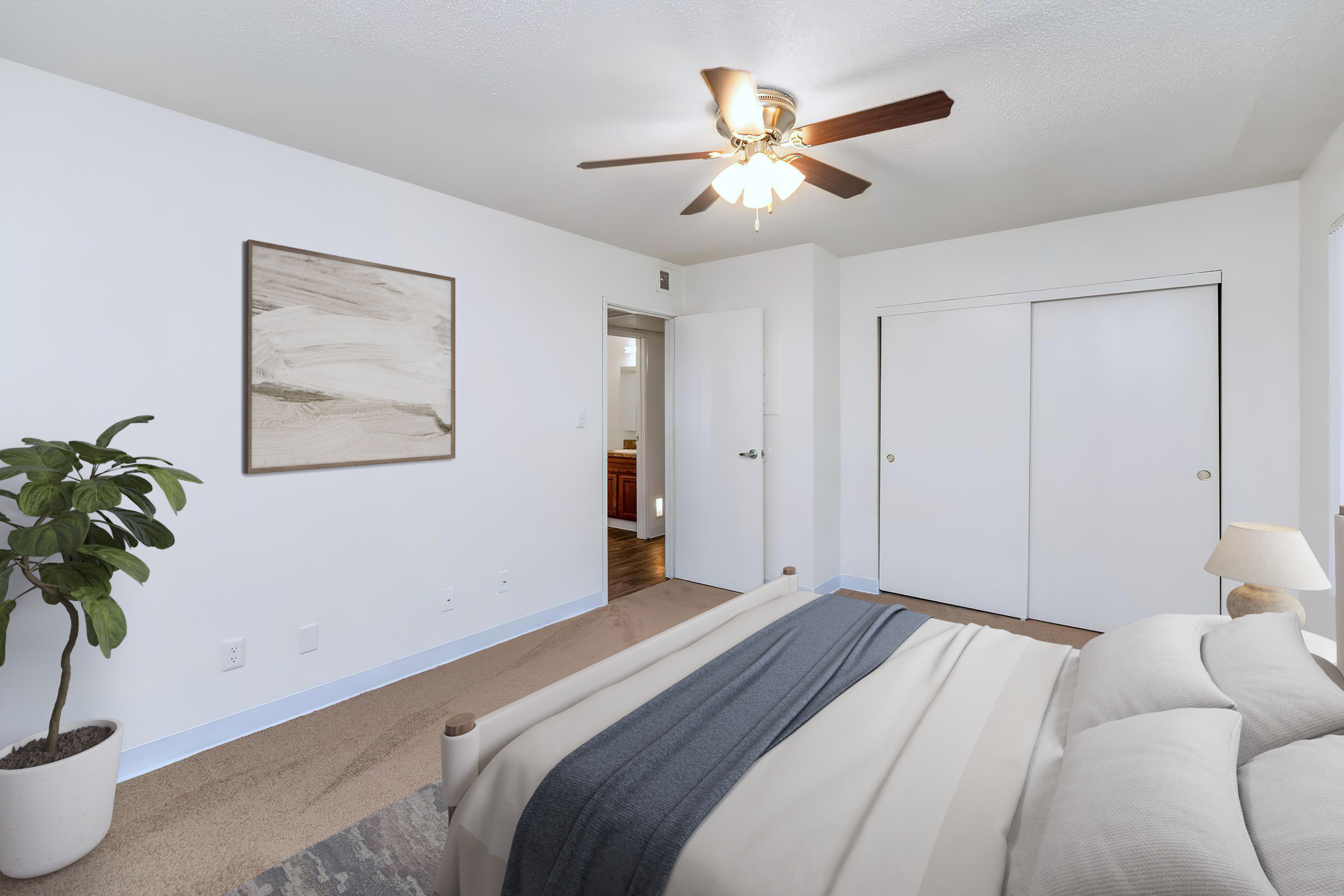 A well-lit bedroom featuring a queen-sized bed with white bedding and a gray throw blanket. A ceiling fan and a green potted plant add to the decor. There's a piece of abstract artwork on the wall, a light lamp on a bedside table, and closed sliding closet doors leading to a bathroom. The walls are painted white, creating a clean and spacious feel.