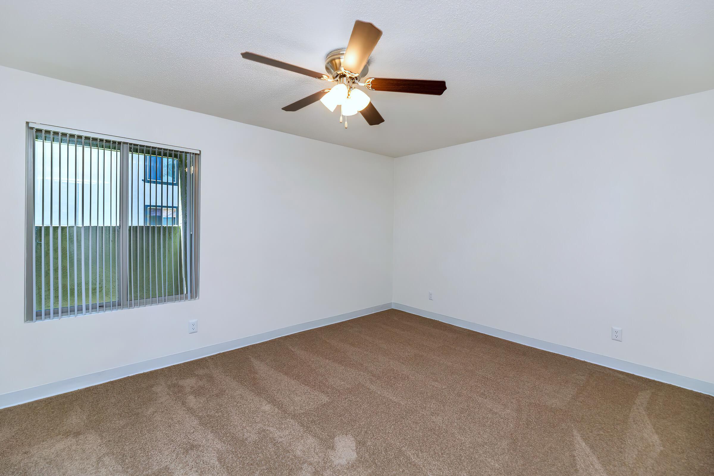 A spacious, empty room featuring light beige carpet flooring, a ceiling fan with four light bulbs, and a large window with vertical blinds allowing natural light. The walls are painted white, creating a bright and open atmosphere.