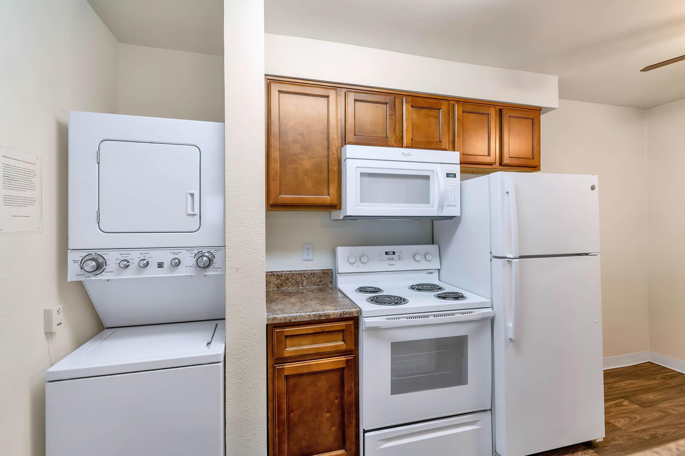 A compact laundry area with a stacked washer and dryer on the left, next to a kitchen featuring brown cabinetry, a white microwave, an electric stove, and a full-sized refrigerator. The kitchen has a neutral-colored wall and wooden flooring, creating a practical and modern space.