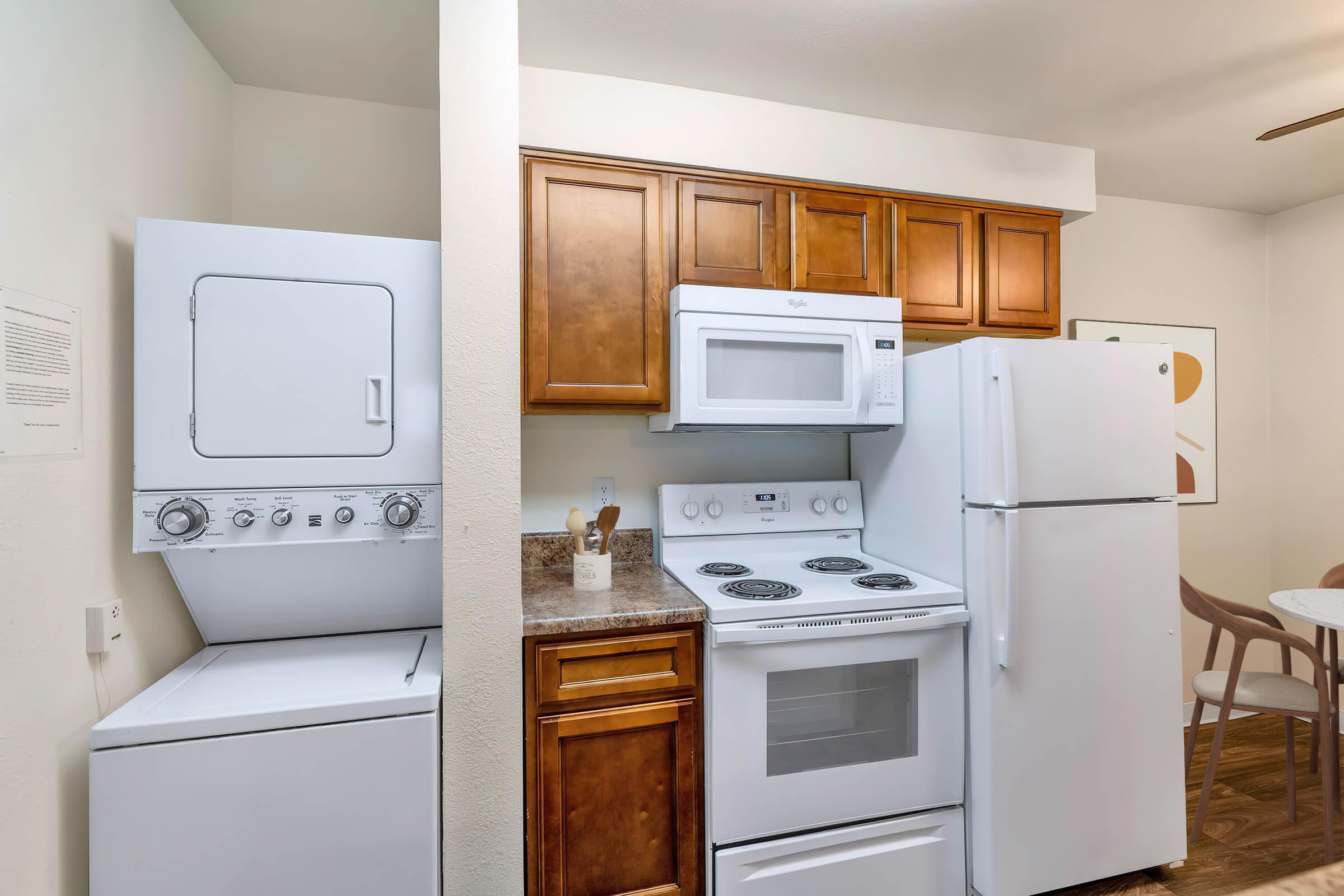 Compact kitchen featuring a stacked washer and dryer on the left, followed by a microwave, oven, and a refrigerator with wooden cabinets above them. Light-colored walls enhance the bright and clean aesthetic, with a dining table visible in the background.