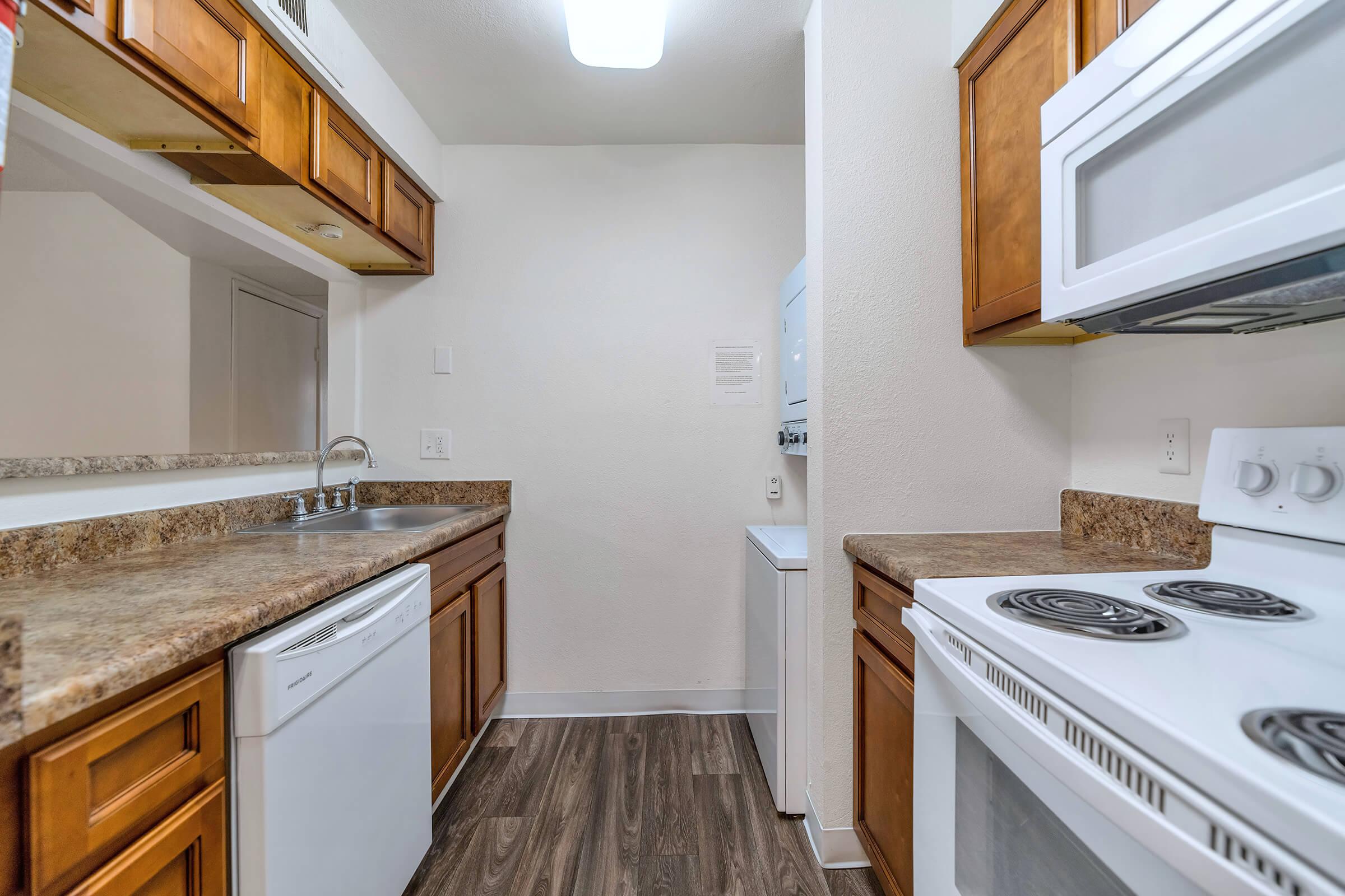 A clean and modern kitchen featuring wooden cabinets, a stainless steel sink, a dishwasher, and a white stove with an oven. The countertop is a dark, speckled surface. A washing machine is visible in the corner, and the walls are painted a light color, creating a bright and spacious atmosphere.