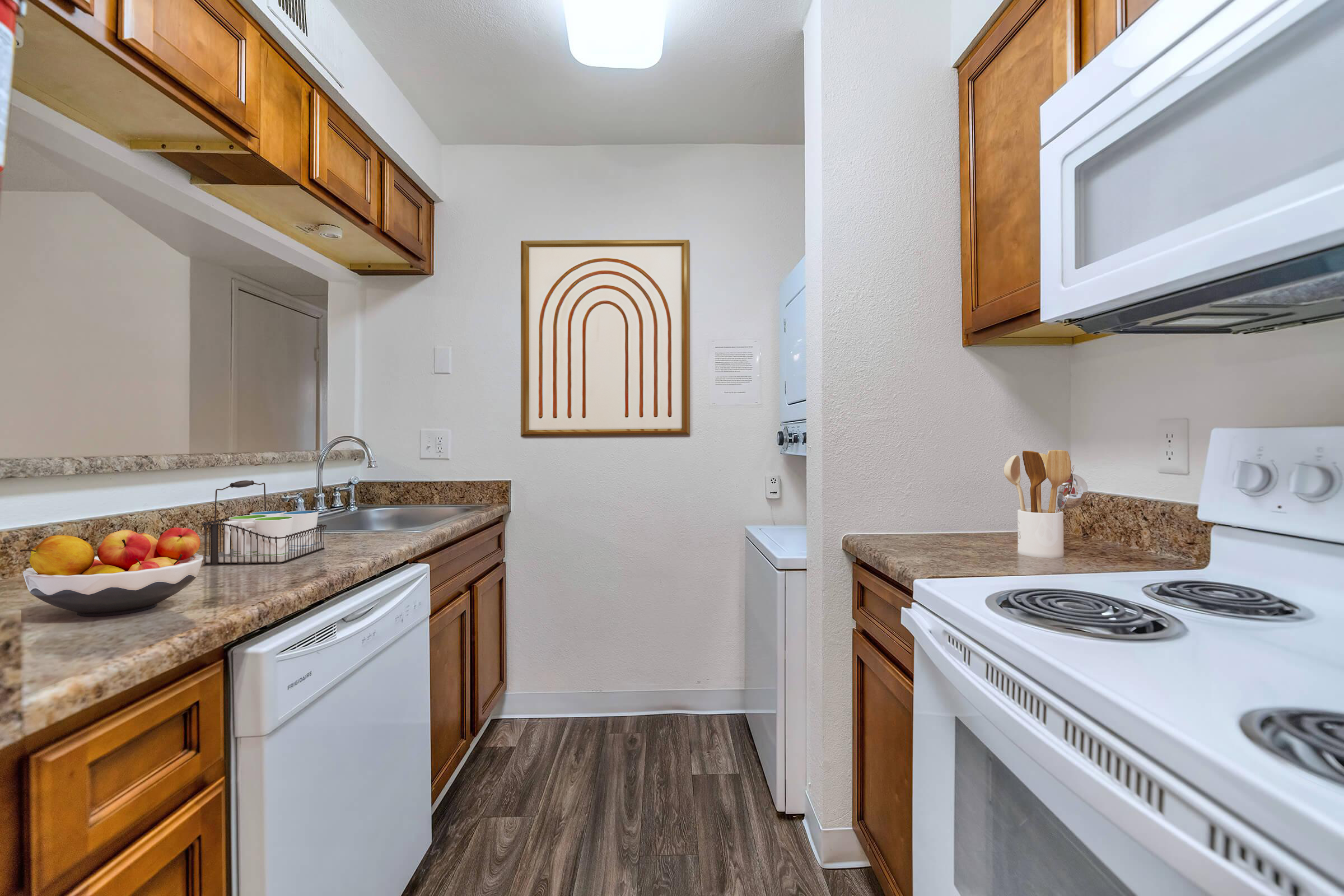 A modern kitchen featuring wood cabinets, a granite countertop, and stainless steel appliances. There is a bowl of apples on the counter, a washing machine in the corner, and a wall art piece with a rainbow design. The space is well-lit and neatly arranged.