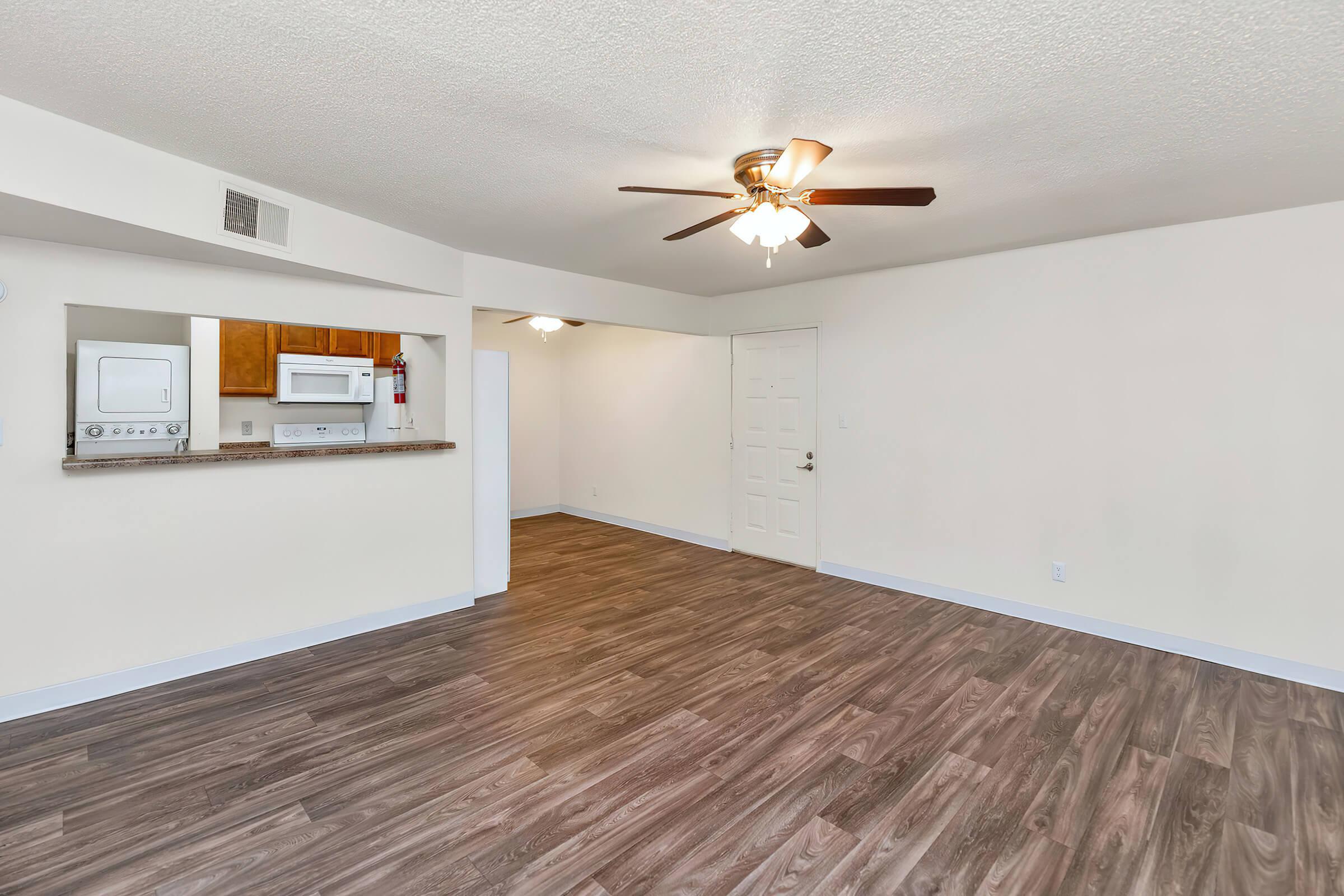 Spacious living area featuring wood-style flooring, a ceiling fan, and natural light. In the background, there is a partial view of a kitchen with light wood cabinets, a microwave, and a washer and dryer unit. The walls are painted in a soft, neutral color.