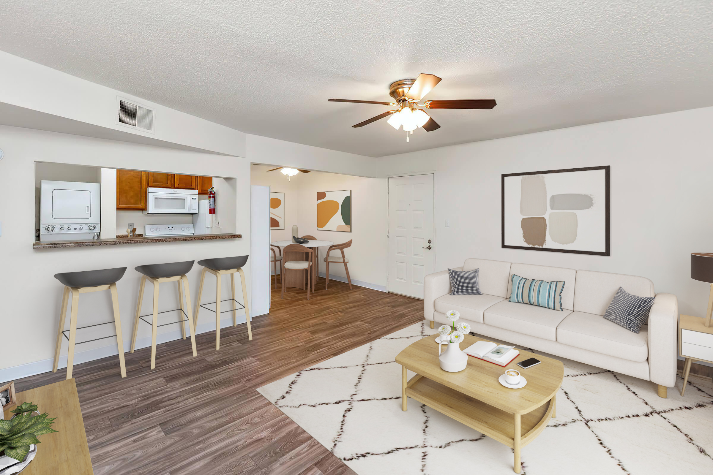 A modern living room and kitchen area featuring a light-colored sofa, a wooden coffee table, and stylish bar stools. The decor includes artwork on the walls, a ceiling fan, and wood-style flooring. A microwave and stove are visible in the kitchen area, creating a cozy, inviting space.