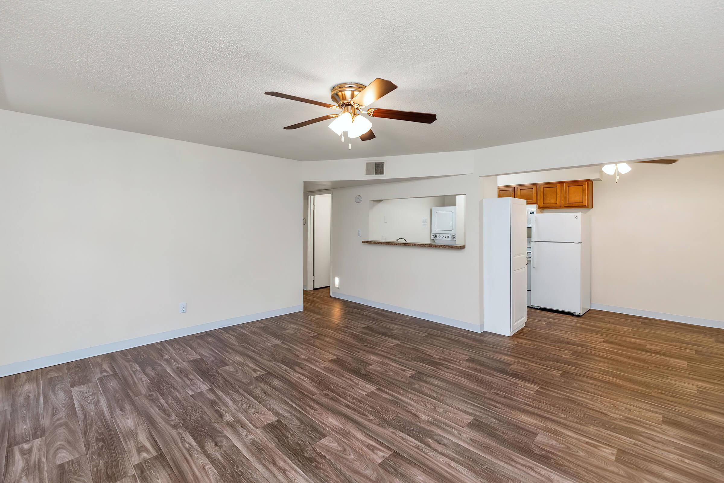 A spacious, well-lit living area with a ceiling fan, featuring light-colored walls and dark wood laminate flooring. The open layout leads to a kitchen area with wooden cabinetry and a white refrigerator visible in the background. Natural light filters in from adjacent rooms.