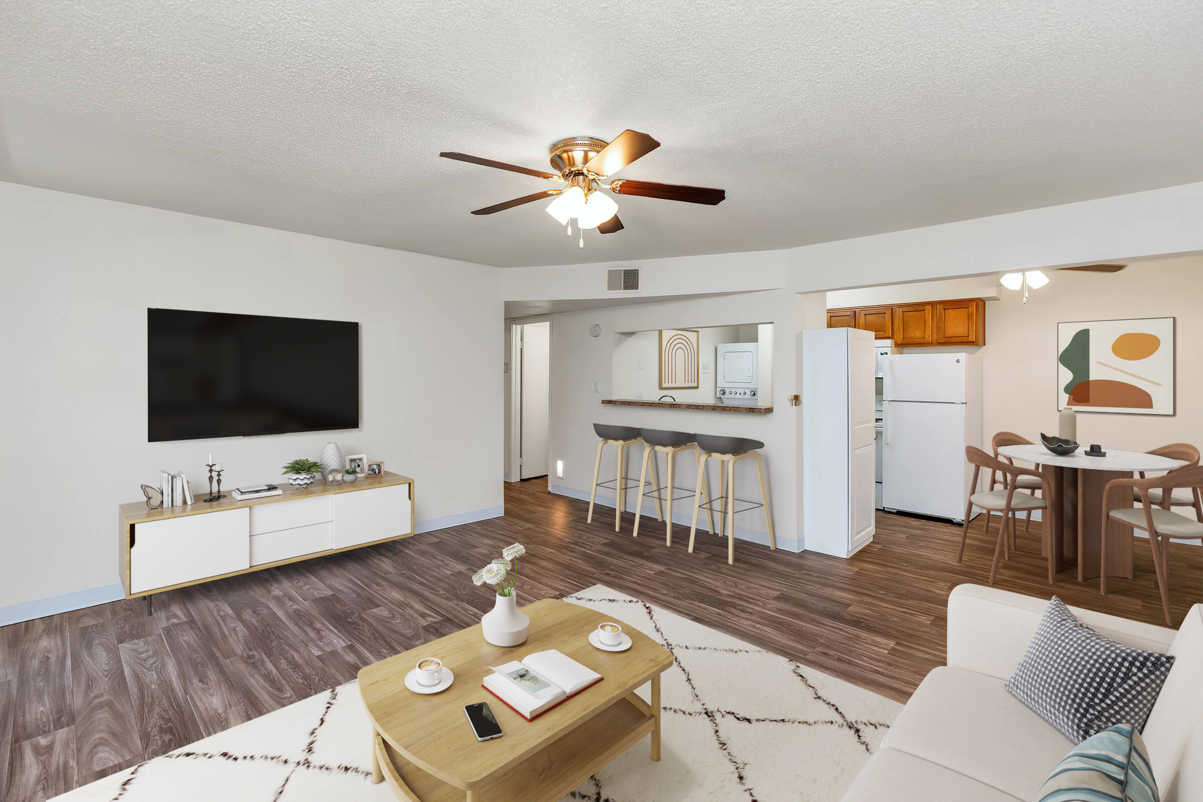 A bright, modern living room featuring a ceiling fan, a wall-mounted television, and a beige sofa. A wooden coffee table with books and a plant sits in the center. Two barstools are positioned at a kitchen counter in the background, adjacent to a dining area with a small table and four chairs.
