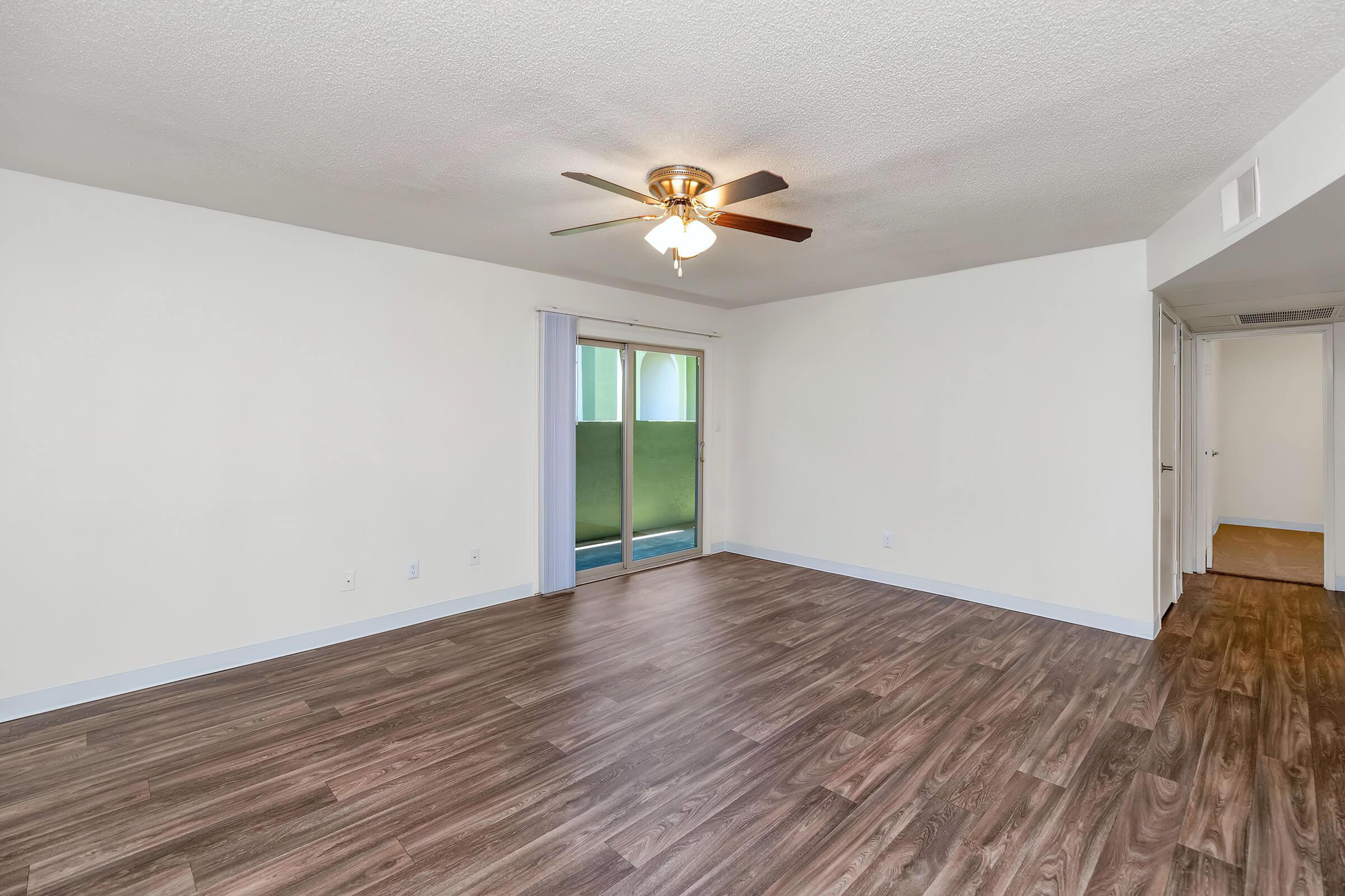 A spacious living room with a ceiling fan, light wood-like flooring, and neutral-colored walls. There is a sliding glass door that leads to a balcony or patio area. The room has a bright and open feel, with one corner showing a doorway leading to another room. Natural light streams in through the glass door.