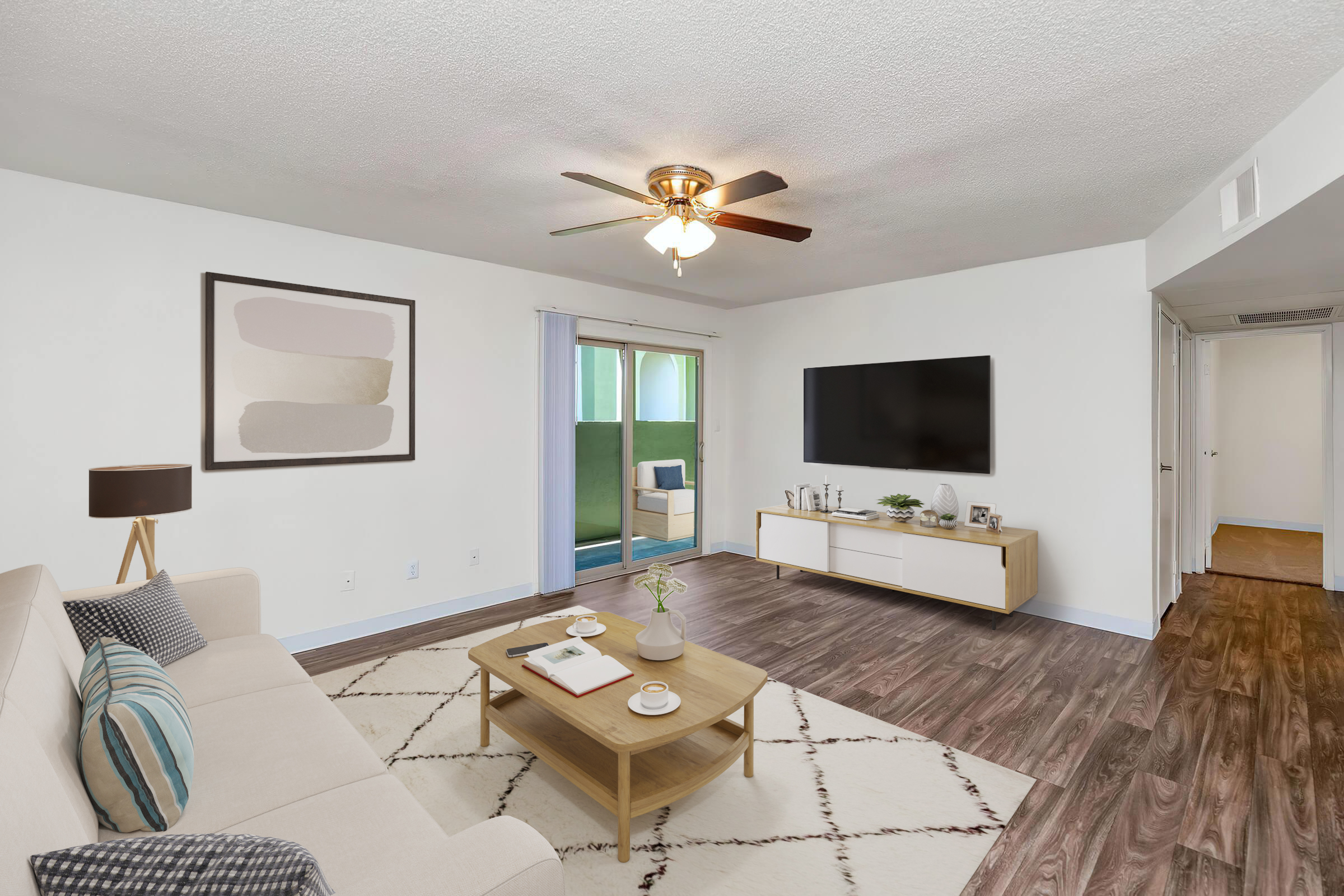 A modern living room featuring a light-colored sofa with decorative cushions, a coffee table with books and a plant, and a large TV mounted on the wall. The space has a ceiling fan, a piece of abstract wall art, and a door leading to a balcony, with warm hardwood-style flooring throughout.