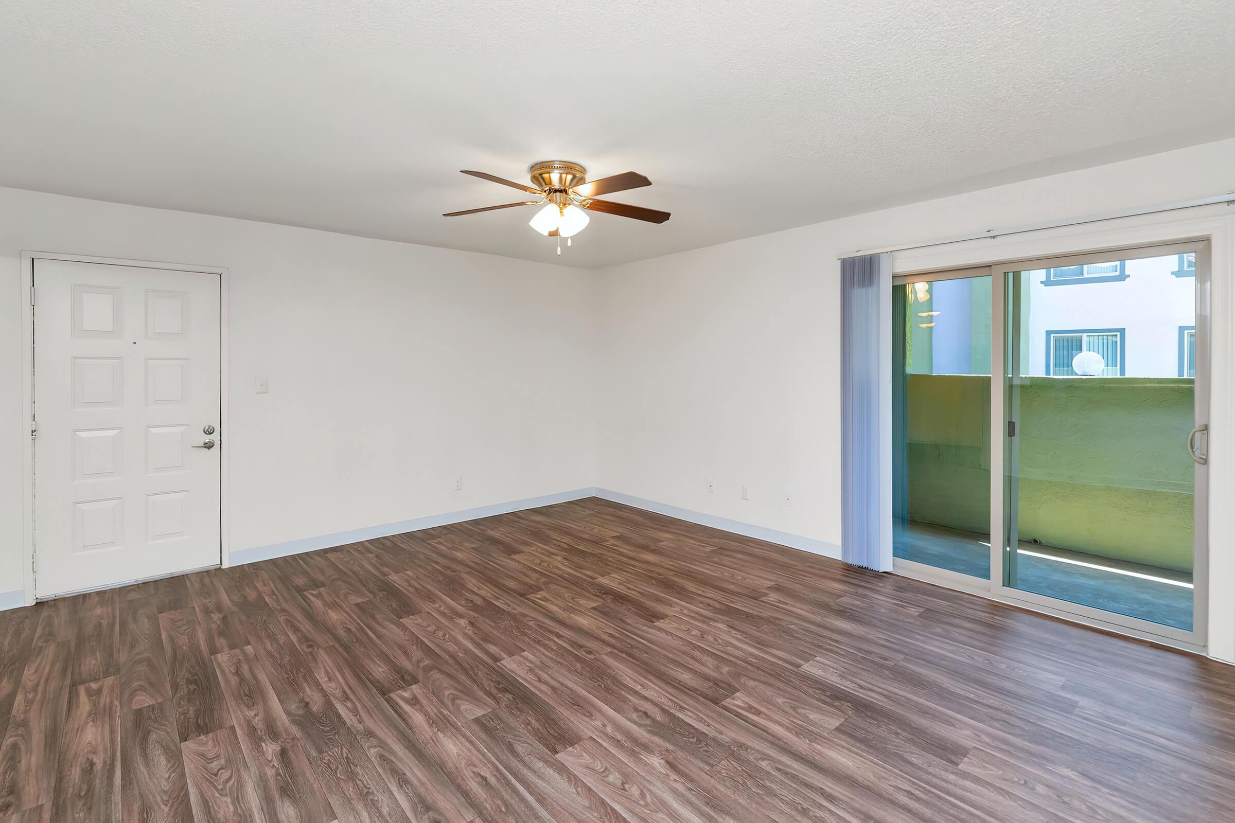 A spacious, empty living room with light-colored walls and a ceiling fan. The floor features dark wood-like laminate. To the right, there is a sliding glass door leading to a small outdoor area, while a plain white door is visible on the left side of the room. Natural light fills the space.