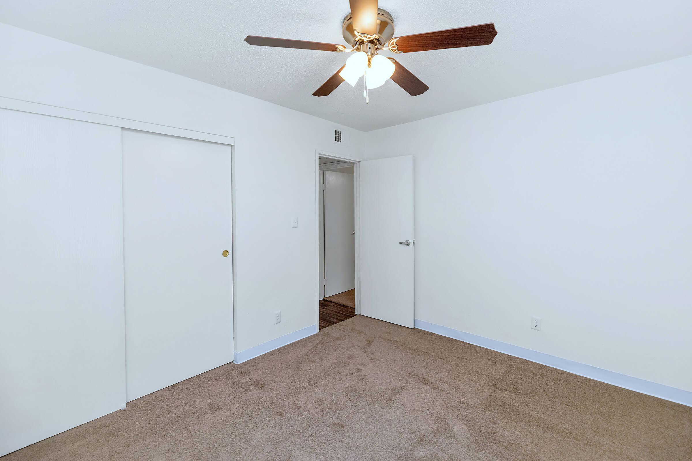 A small, empty room with beige carpet and white walls. A ceiling fan with wooden blades hangs from the ceiling. There are two doors: one closet door on the left and an open door leading to another room on the right. Natural light brightens the space, creating a clean and inviting atmosphere.