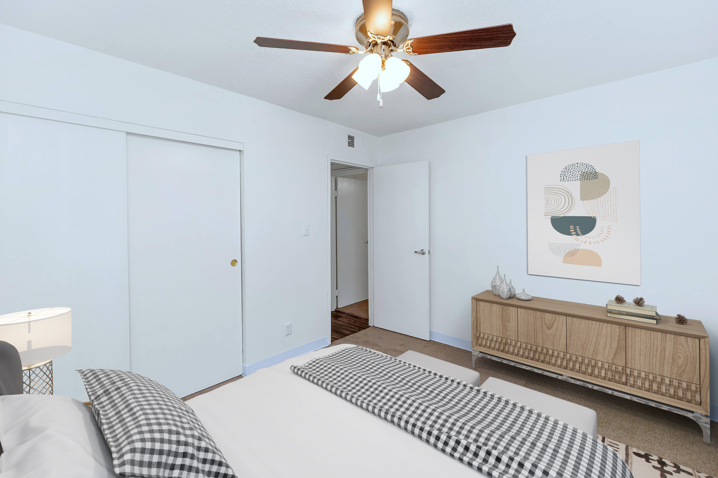 A cozy bedroom featuring a large window, a ceiling fan with wooden blades, and a neatly made bed with a checkered blanket. There is a stylish wooden dresser against the wall, a piece of abstract art hanging nearby, and light blue walls creating a calm ambiance. A closet door and room entrance are also visible.