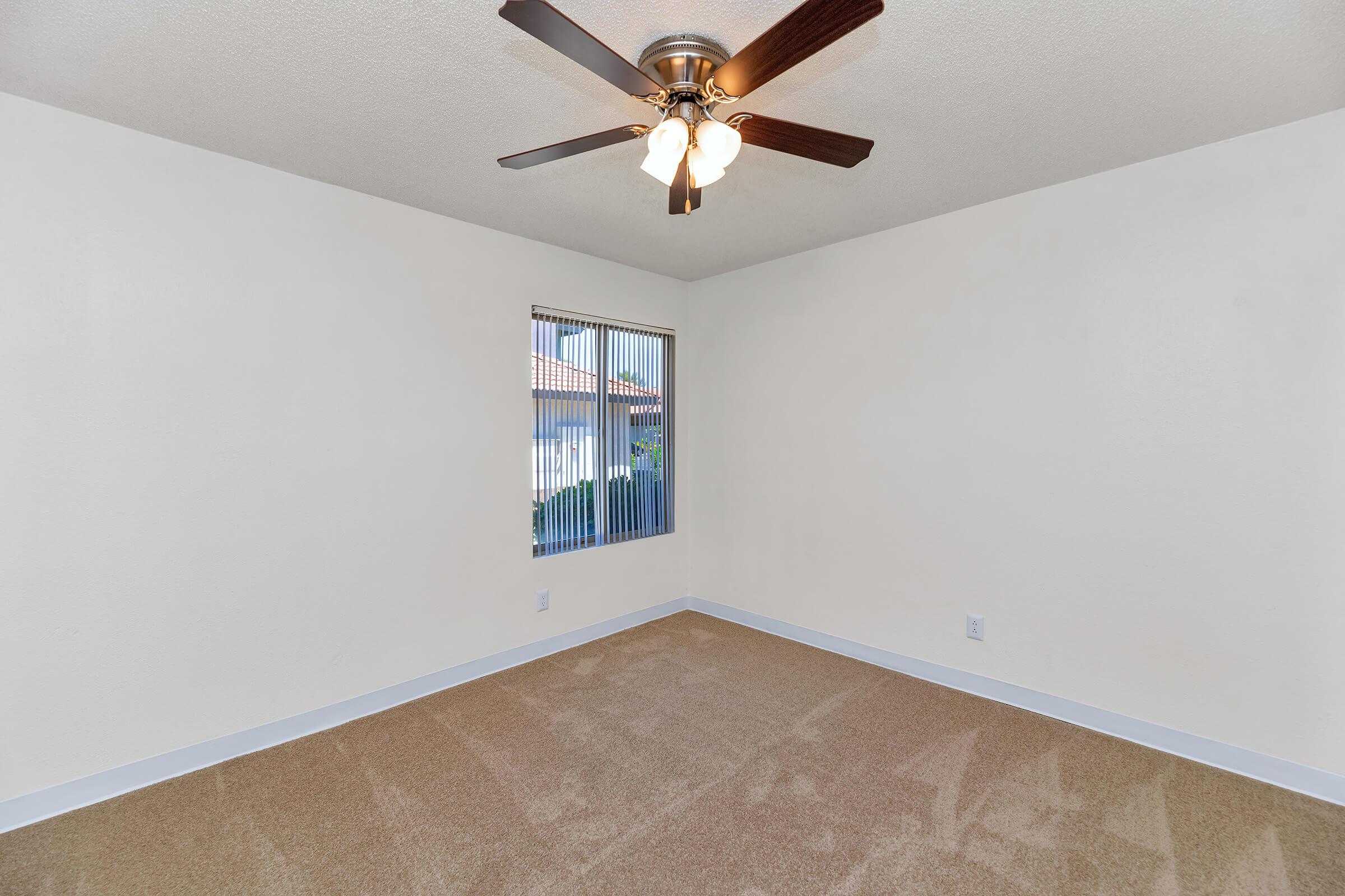 A spacious, well-lit room featuring a ceiling fan with four lights, beige carpet flooring, and a window with vertical blinds. The walls are painted a soft beige color, providing a neutral backdrop. The room appears clean and freshly maintained, suitable for various uses such as a bedroom or office.