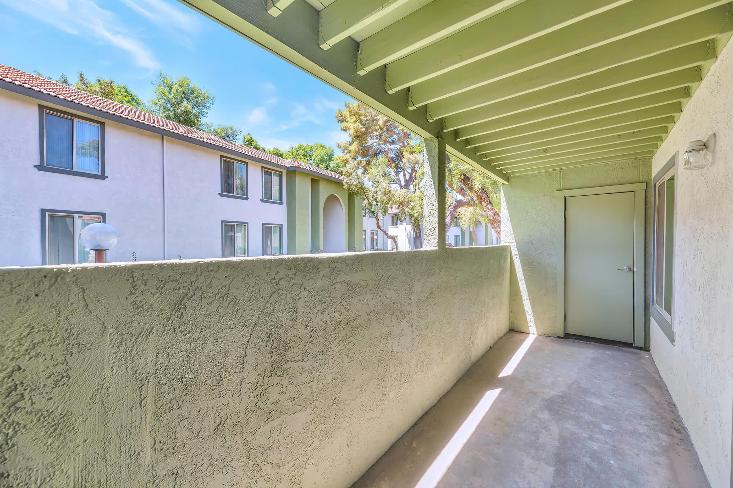 Empty balcony area with a concrete floor and light green beams overhead. The view includes neighboring apartment buildings surrounded by trees under a clear blue sky. A door leads to a room, and a round light fixture is visible on the wall. Sunlight casts soft shadows on the surface.