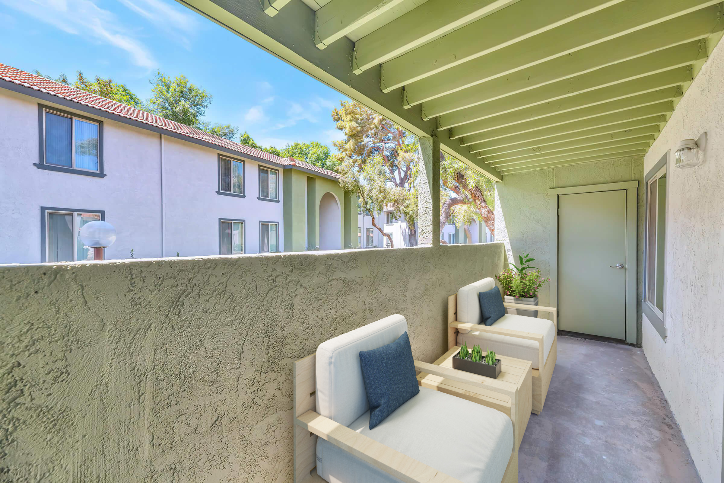 A balcony with two modern white chairs and a small table, adorned with a potted plant. It's surrounded by a textured concrete wall and overlooks an apartment complex with trees in the background, under a clear blue sky. The area is well-lit and inviting, suggesting a comfortable outdoor space.