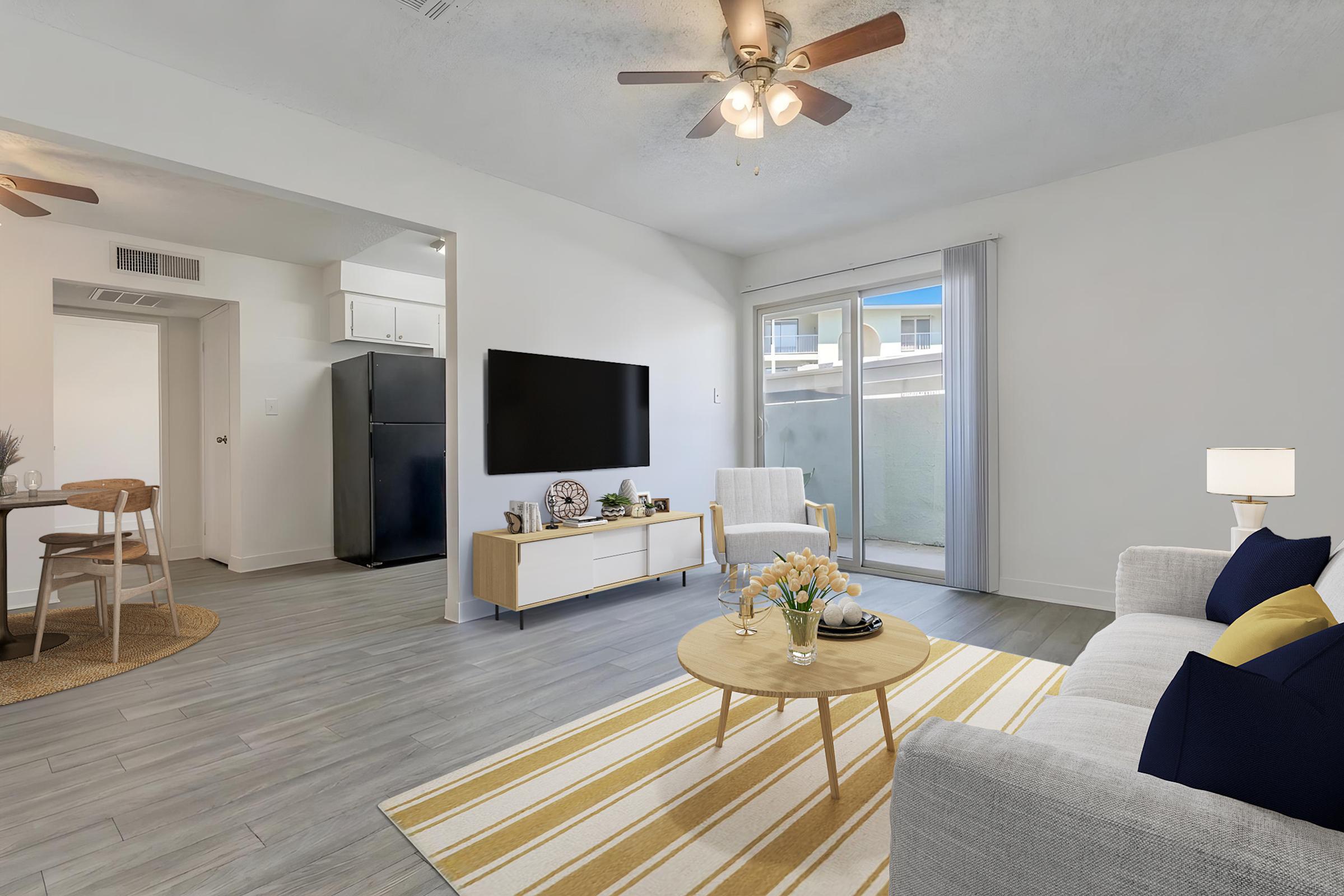 A modern living room featuring a light gray sofa with blue and yellow cushions, a round coffee table with flowers, and a television mounted on the wall. There are two armchairs and a small dining area in the background, with a kitchen visible. Large windows provide natural light.