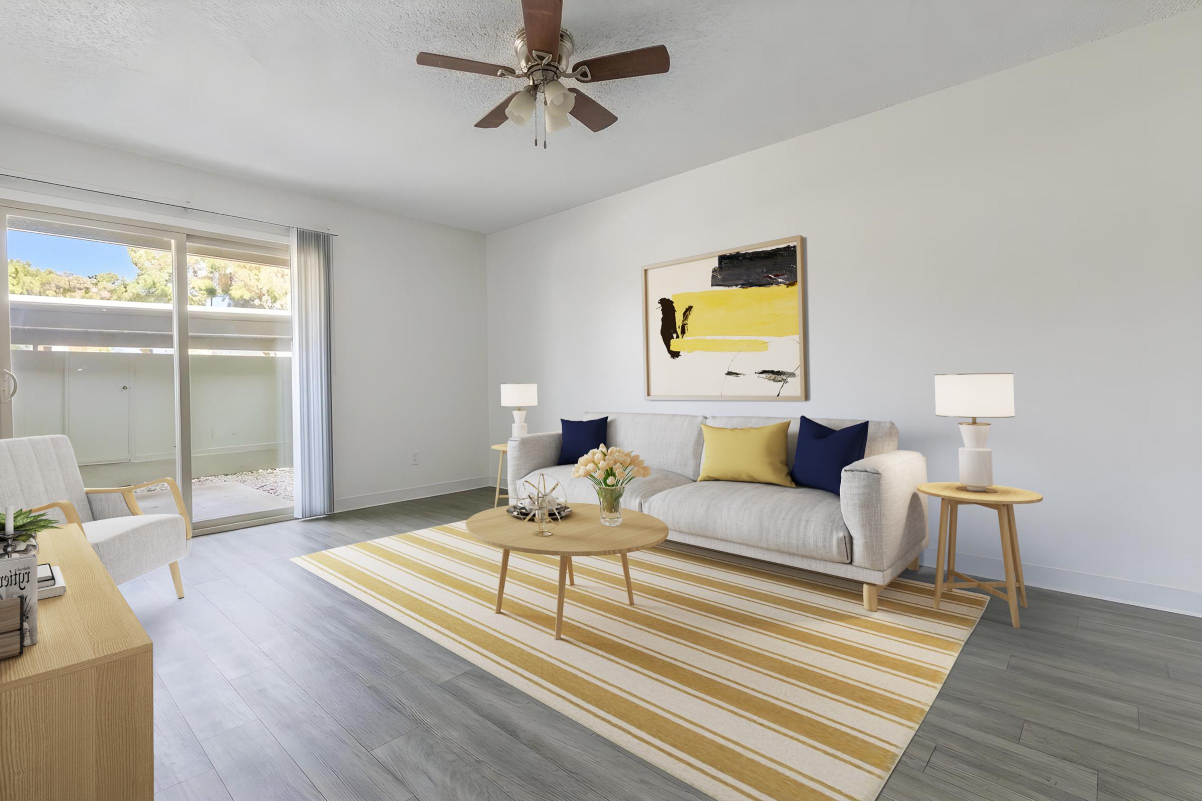 Bright and airy living room featuring a light-colored sofa with blue and yellow pillows, a round coffee table with a flower vase, and decorative items. A large sliding glass door provides natural light, revealing an outdoor area. The floor has light gray planks and a striped yellow and white area rug.