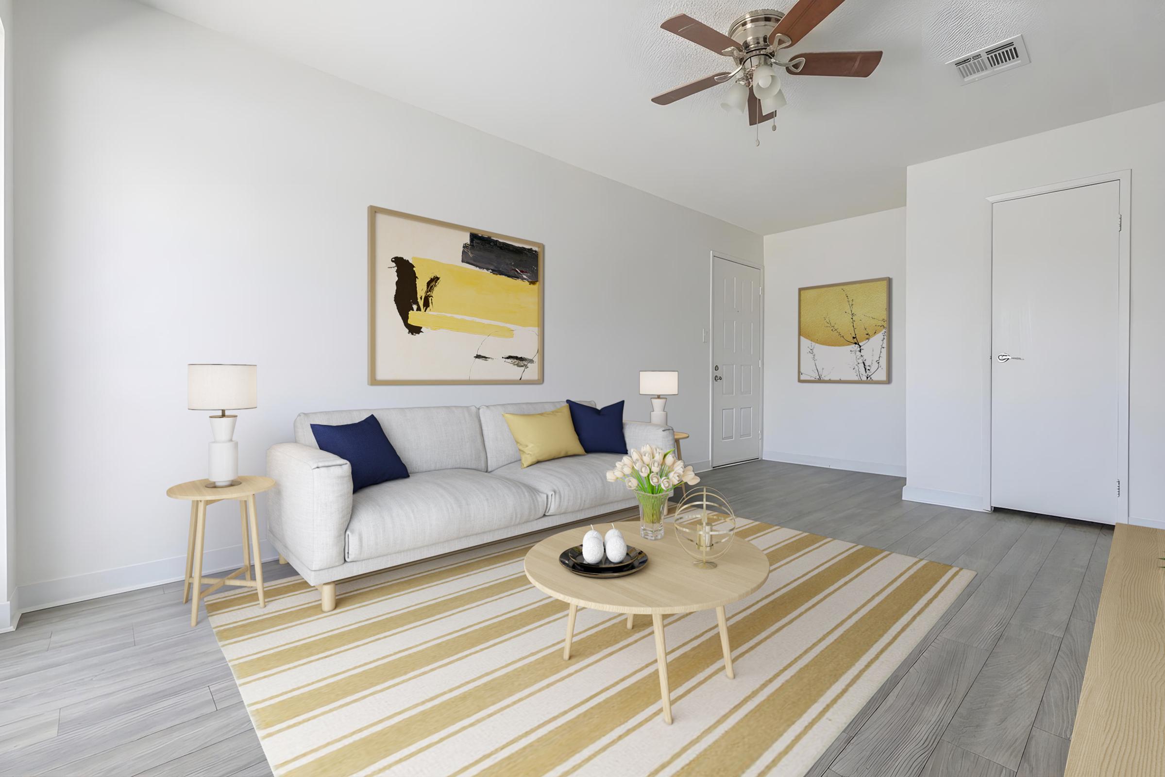 A bright and airy living room featuring a light-colored sofa with blue and yellow cushions, a round coffee table, and decorative items including flowers and candles. The floor is covered with a striped rug, and there are two framed abstract paintings on the walls. A ceiling fan is visible above.