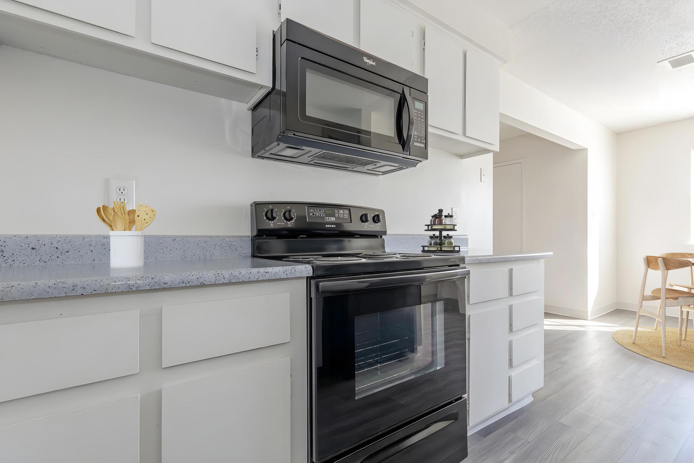 Modern kitchen featuring a black microwave and stove, with light gray countertops. White cabinets provide storage, and a small decorative item on the counter adds a touch of style. The kitchen has a bright, airy feel with natural light coming from the adjacent room.