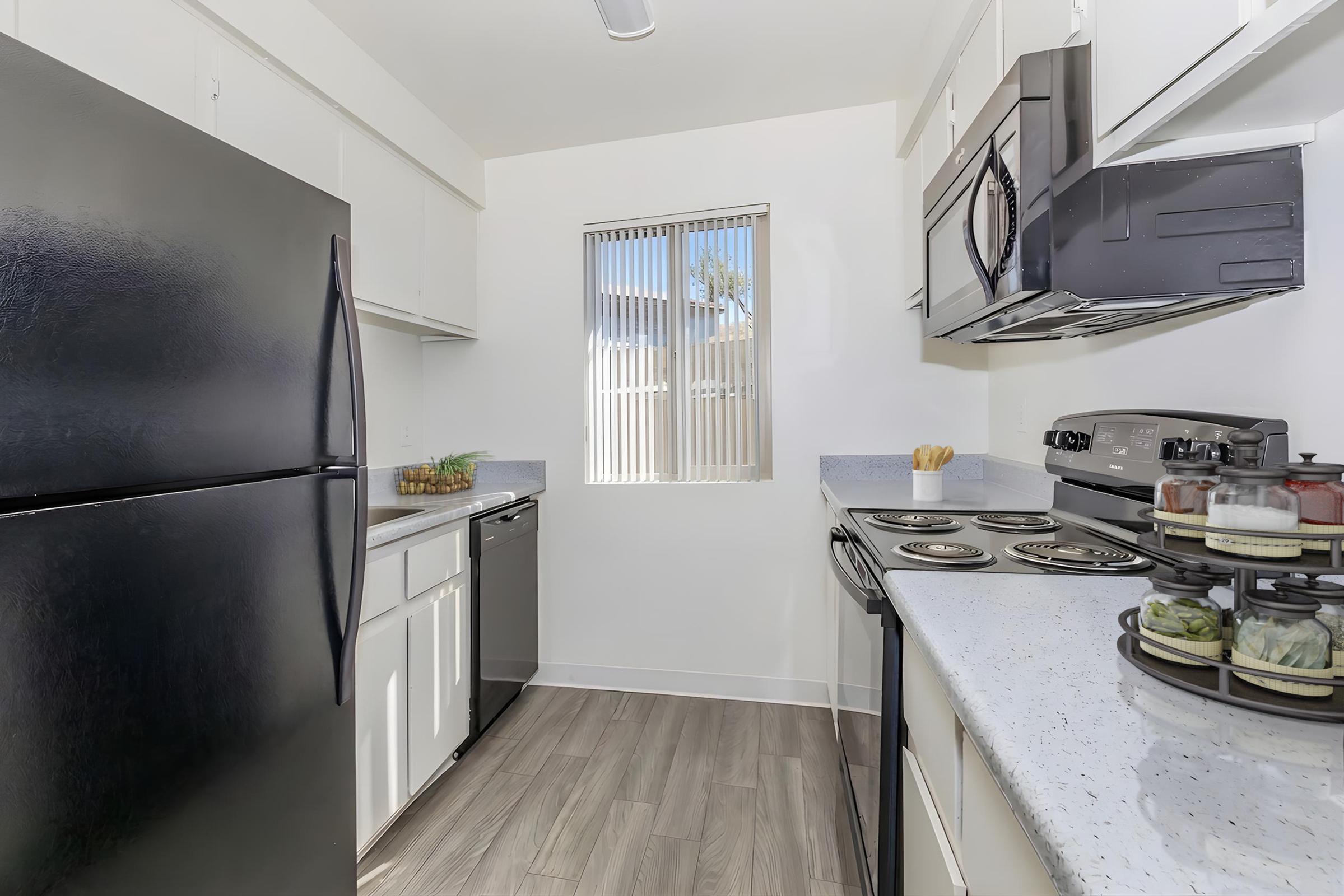 Modern kitchen featuring black appliances, including a refrigerator, stove, and microwave. White cabinetry contrasts with the dark appliances, and a window allows natural light to enter. The countertops are light-colored, and there is a glimpse of organized storage near the stove.