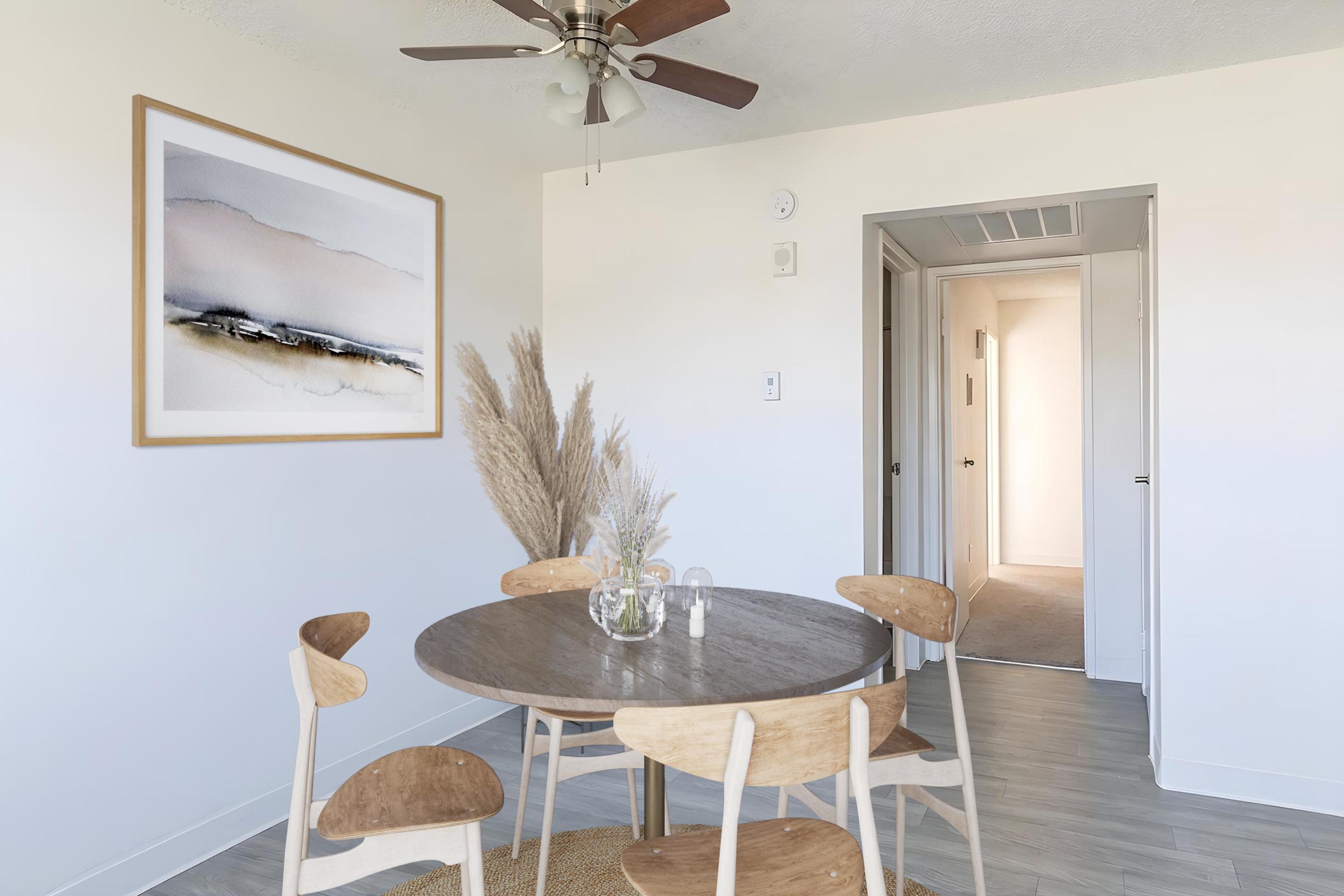 A modern dining area featuring a round wooden table surrounded by four wooden chairs. A large framed abstract artwork hangs on the wall, and a soft area rug is beneath the table. The space is bright and airy, with a ceiling fan and a doorway leading to an adjacent room.