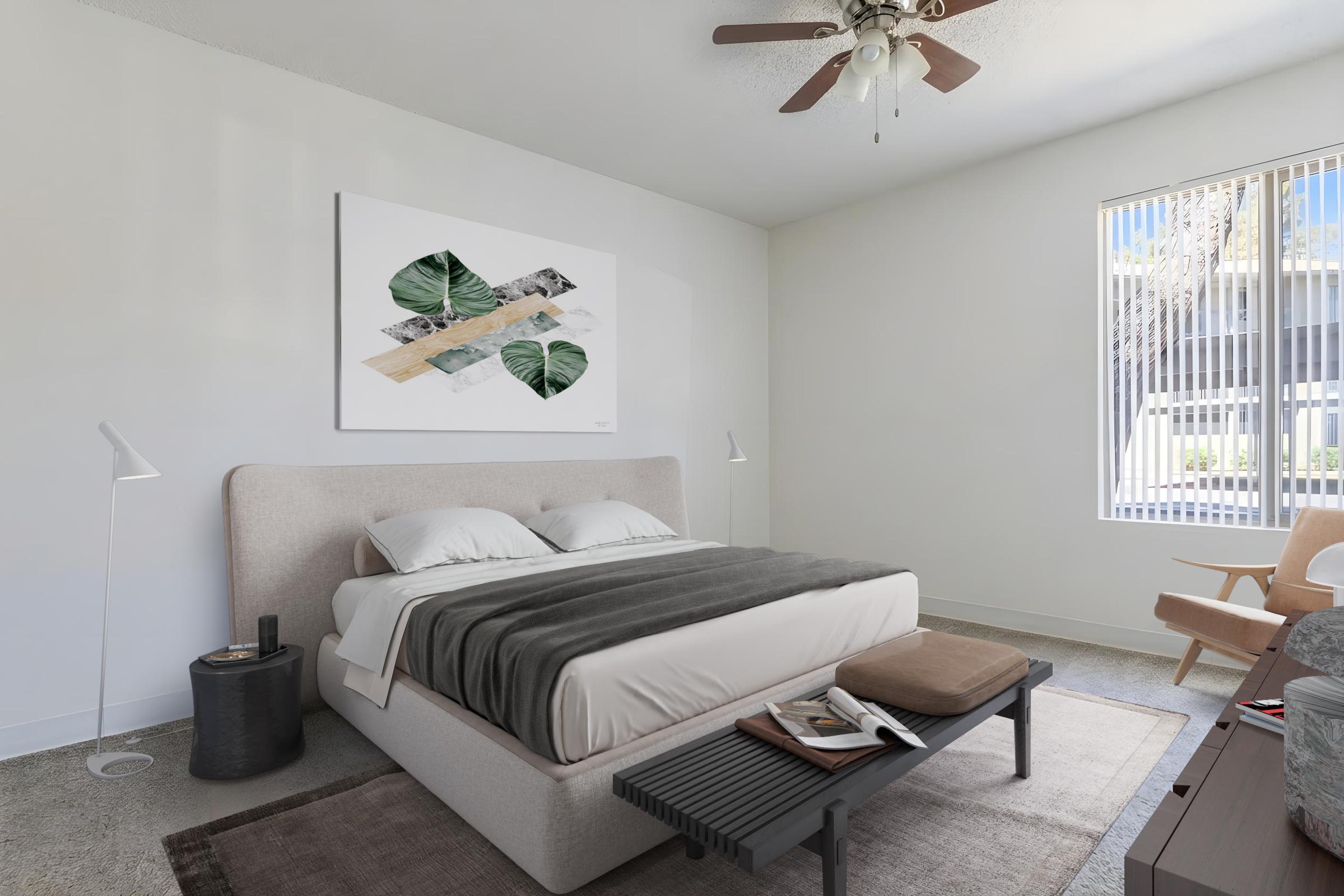 A modern bedroom featuring a large bed with white linens and a gray throw blanket. There’s a painting with leafy designs on the wall, a stylish wooden bench at the foot of the bed, and a cozy armchair nearby. Natural light filters through a window with blinds, and a ceiling fan is visible.