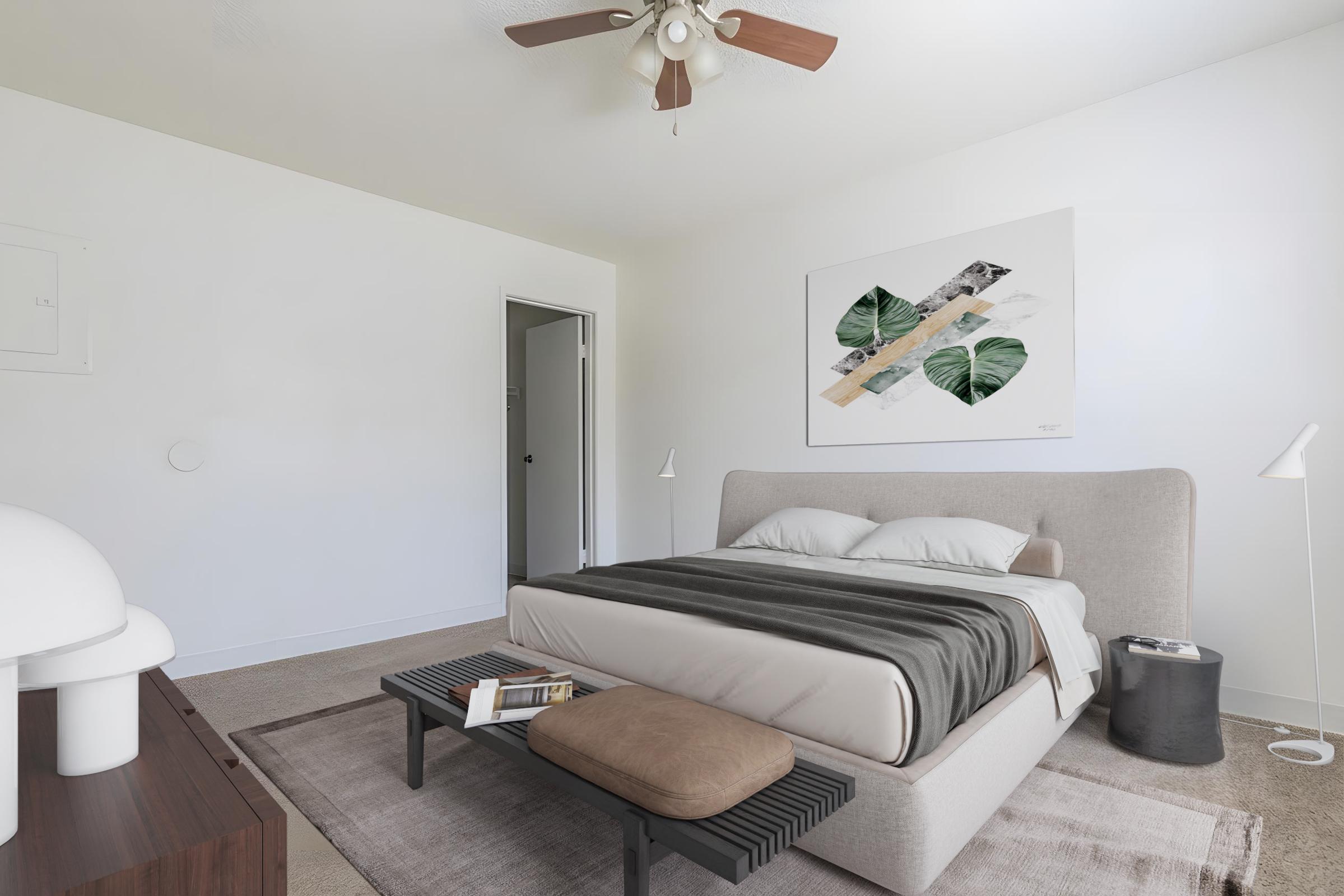 A modern, minimalistic bedroom featuring a bed with a soft gray headboard and white bedding. There is a light-colored rug underneath, a small coffee table with a book, and a sleek lamp. The walls are white, and a piece of abstract wall art with green leaves hangs above the bed. A door leads to another room.