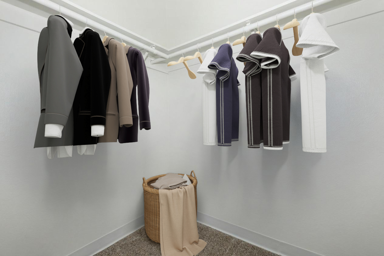 A neatly organized closet featuring a variety of clothing. Several shirts and jackets in neutral colors are hung on wooden hangers, while a wicker laundry basket filled with folded clothes sits on the floor. The walls are plain, and the floor is covered with light-colored carpeting.