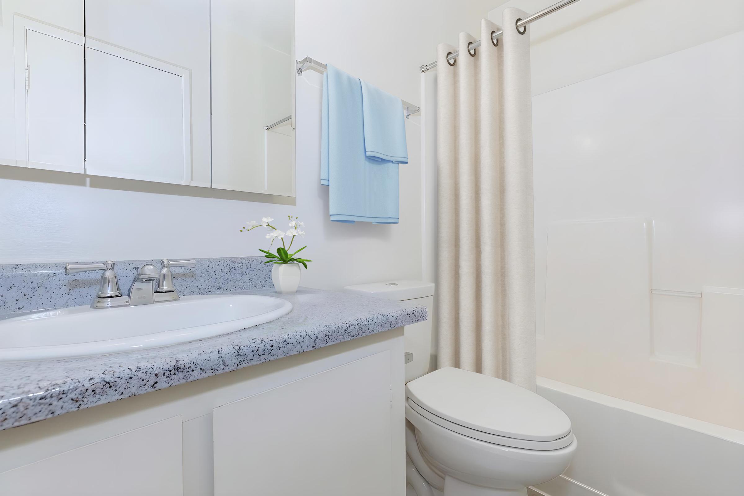 A clean and modern bathroom featuring a white toilet, a sink with a marble countertop, and a shower with a light-colored curtain. A small vase with an orchid adds a touch of decoration, and there are light blue towels hanging on a rack. Mirrors reflect the bright, neutral-colored walls.