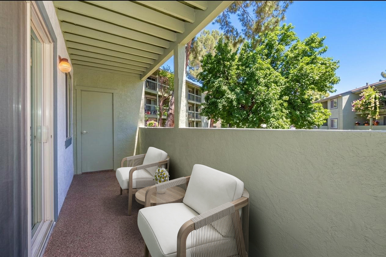 A balcony with two white chairs and a small table, overlooking green trees and a well-maintained courtyard. The setting features soft natural light, enhancing the peaceful ambiance of the outdoor space.