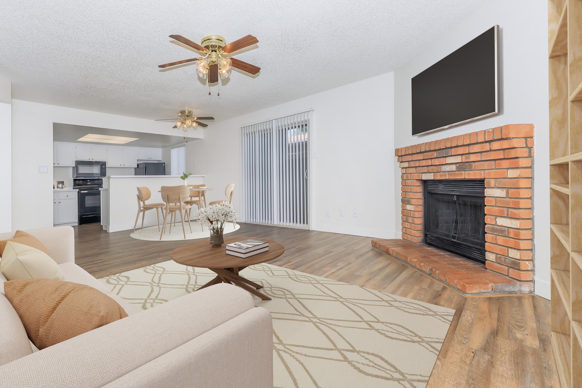 A cozy living room featuring a beige sofa with decorative pillows, a round coffee table, and a brick fireplace. The space has a ceiling fan, light wood flooring, and sliding glass doors leading to an outdoor area. In the background, a dining area with a kitchen is visible, creating an open-concept layout.
