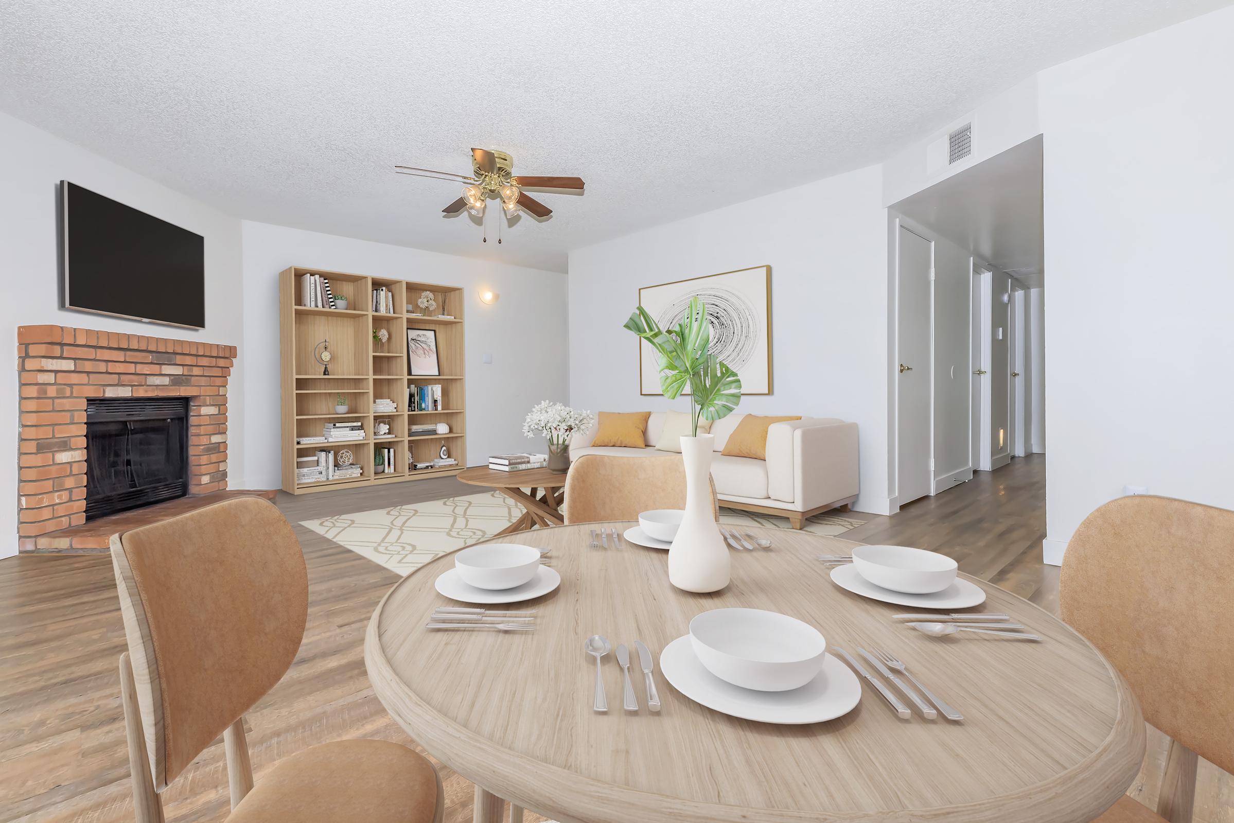 A cozy living room featuring a beige sofa, a coffee table set with white dishes, and a decorative vase. A brick fireplace sits beneath a wall-mounted TV. A wooden bookshelf displays books and decor. Bright overhead lighting and natural light enhance the airy atmosphere. Light wooden flooring complements the warm space.